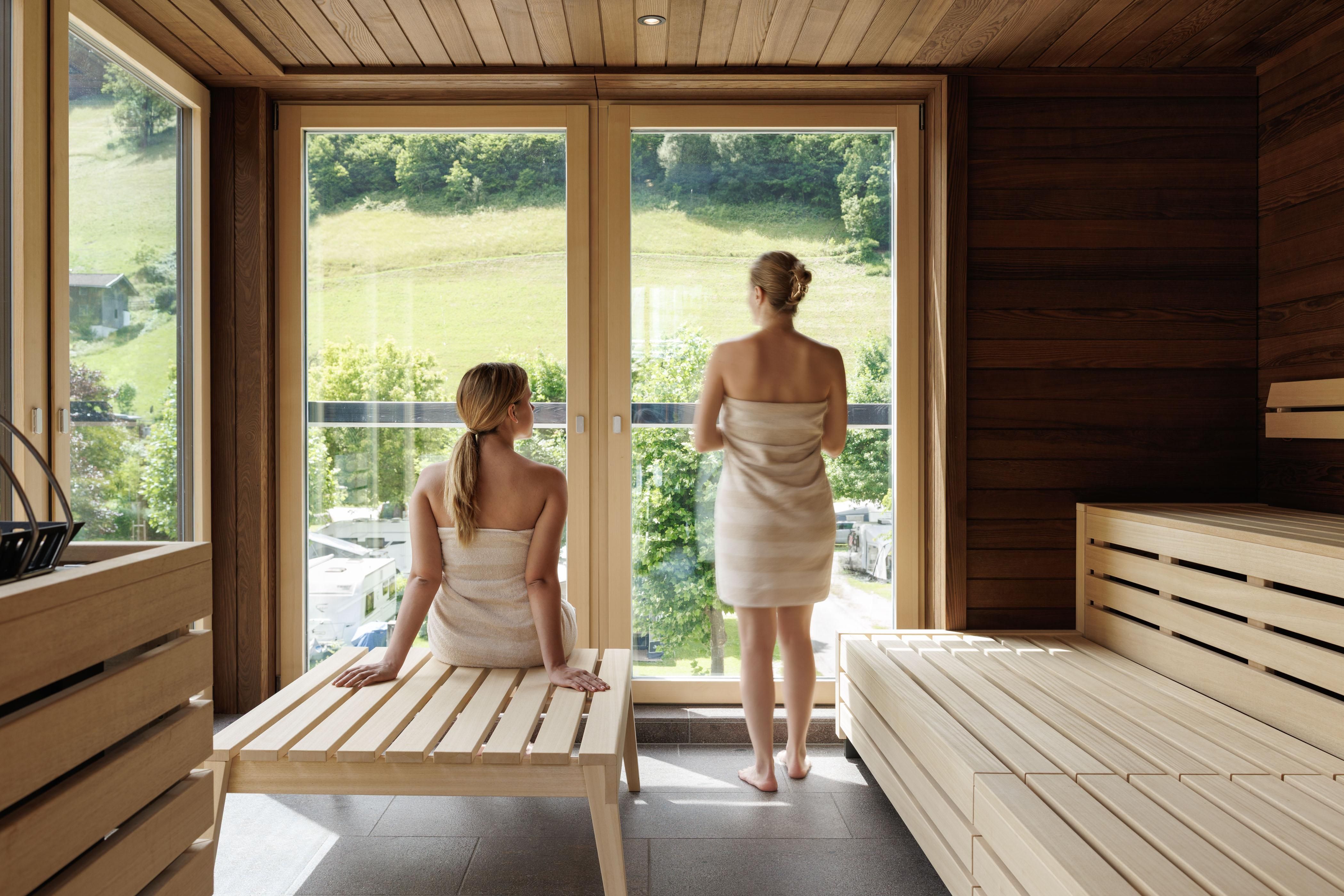Two women in a sauna look out of large windows at the green landscape. The sauna is made of wood and has a relaxing atmosphere.