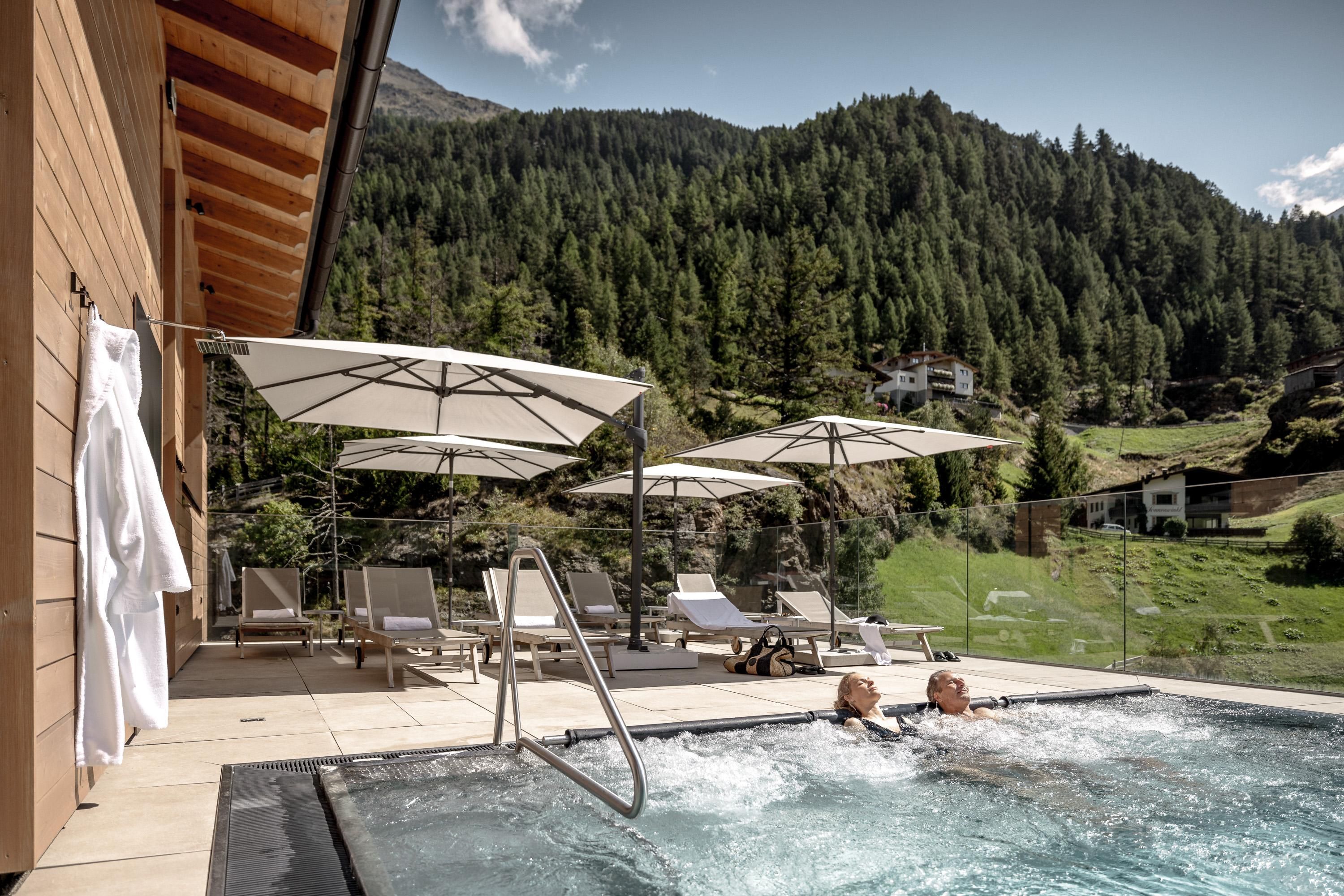 A relaxing outdoor area with a hot tub and loungers. In the background, green mountains and a clear sky can be seen.