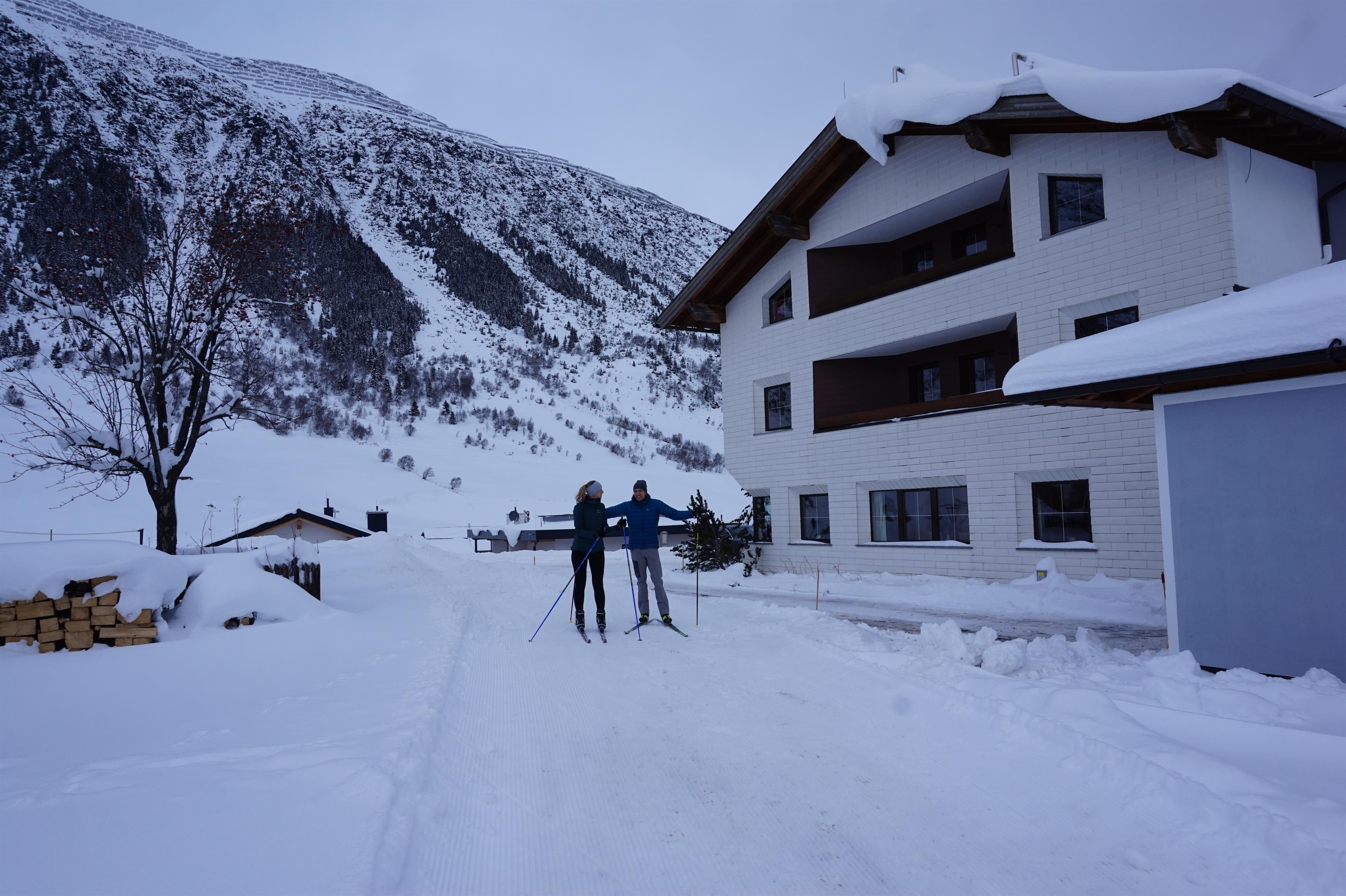 Two skiers are standing on a snow-covered road in front of a white building. Snow-covered mountains can be seen in the background.