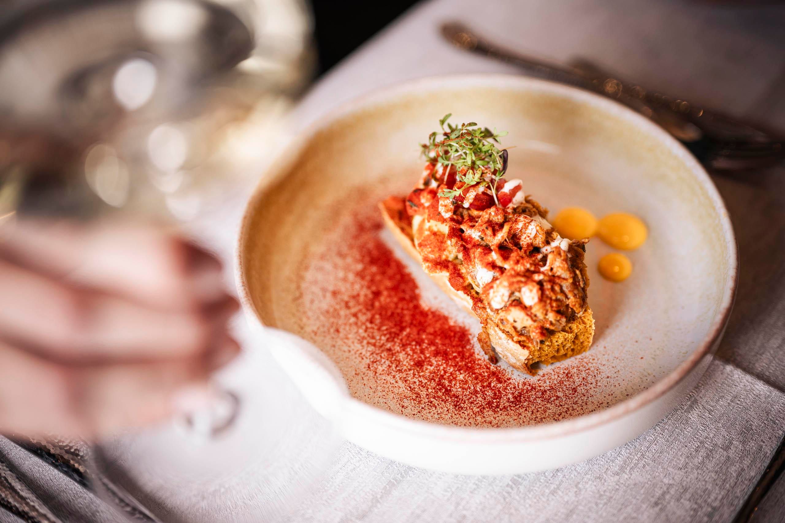 An artfully arranged plate with a piece of fish, garnished with herbs and red spices. In the background, a glass with a drink is visible.