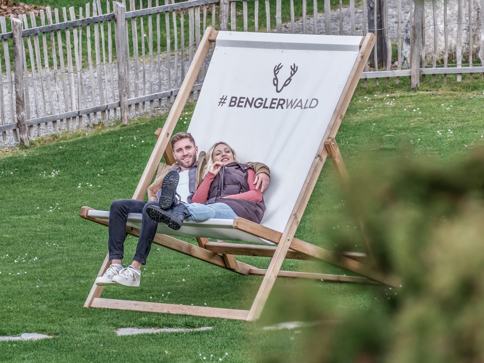 A large deck chair with the inscription "#BENGLERWALD" stands on a green meadow. Two people are relaxing on it and enjoying their time outdoors.