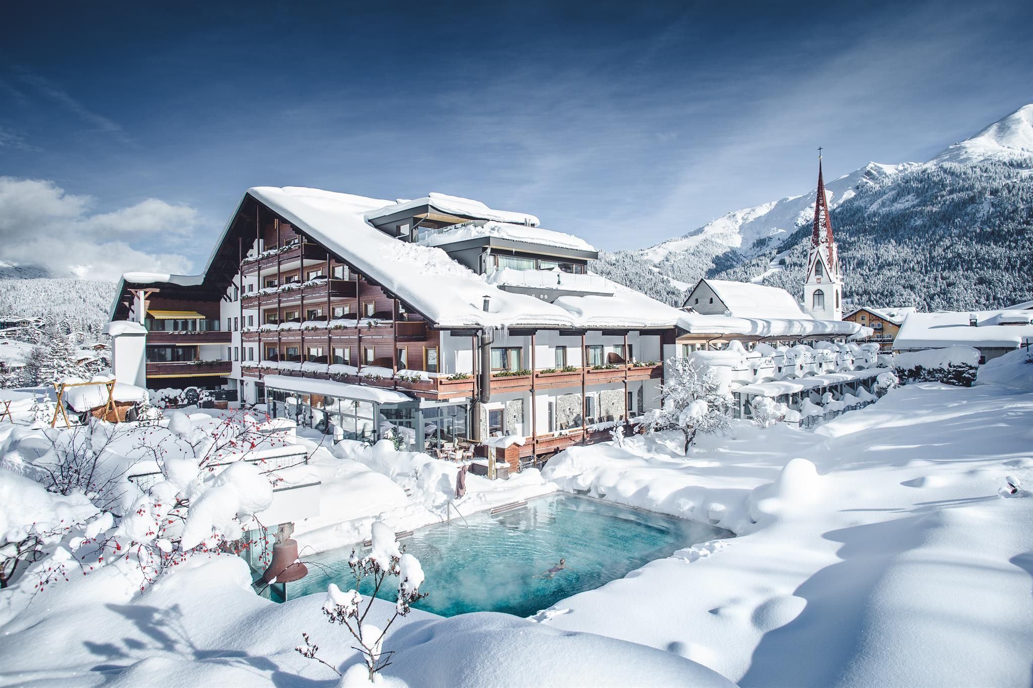 A picturesque hotel in the snow, surrounded by mountains and a clear sky. The garden is covered with fresh snow and the pool adds a touch of coziness.