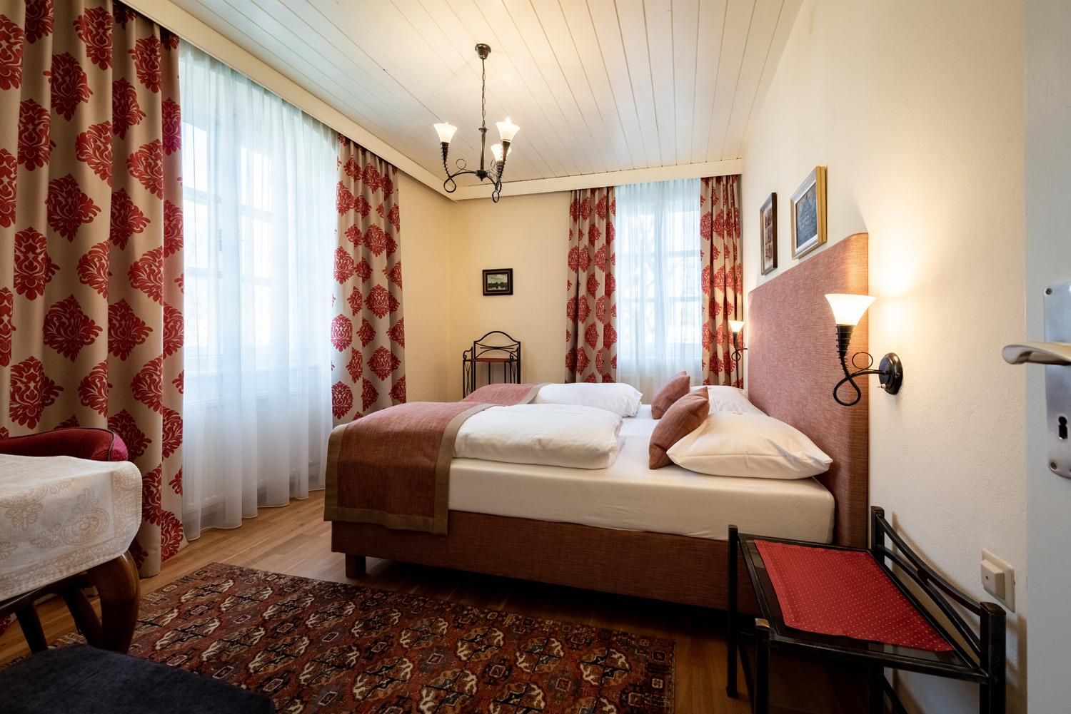 A cozy bedroom with two beds, elegant curtains, and a beautiful rug. The walls are adorned with pictures, and there is warm lighting.