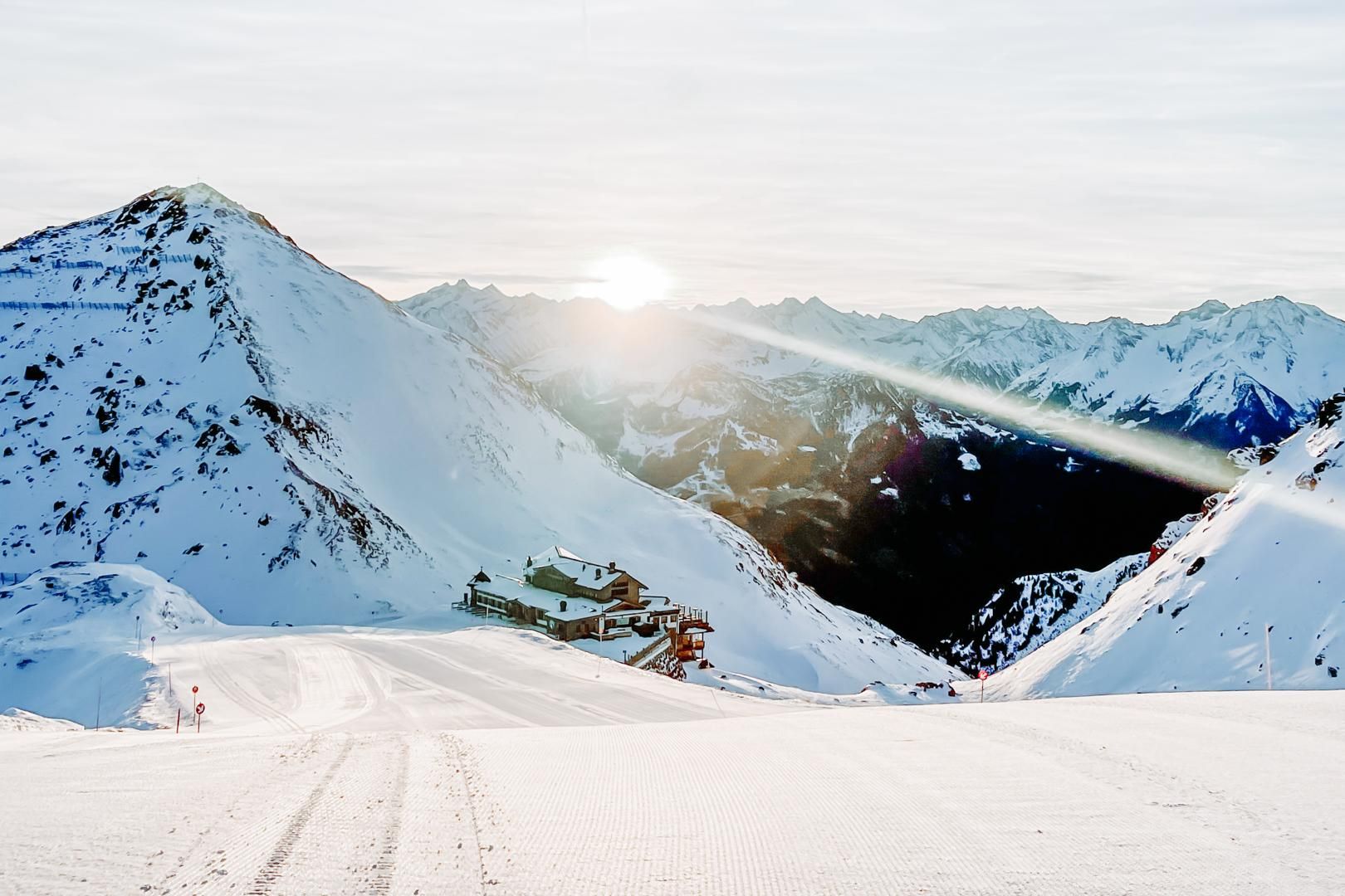 A wintry mountain landscape with snow-covered peaks. The sun shines behind the mountains and brightens the ski run.