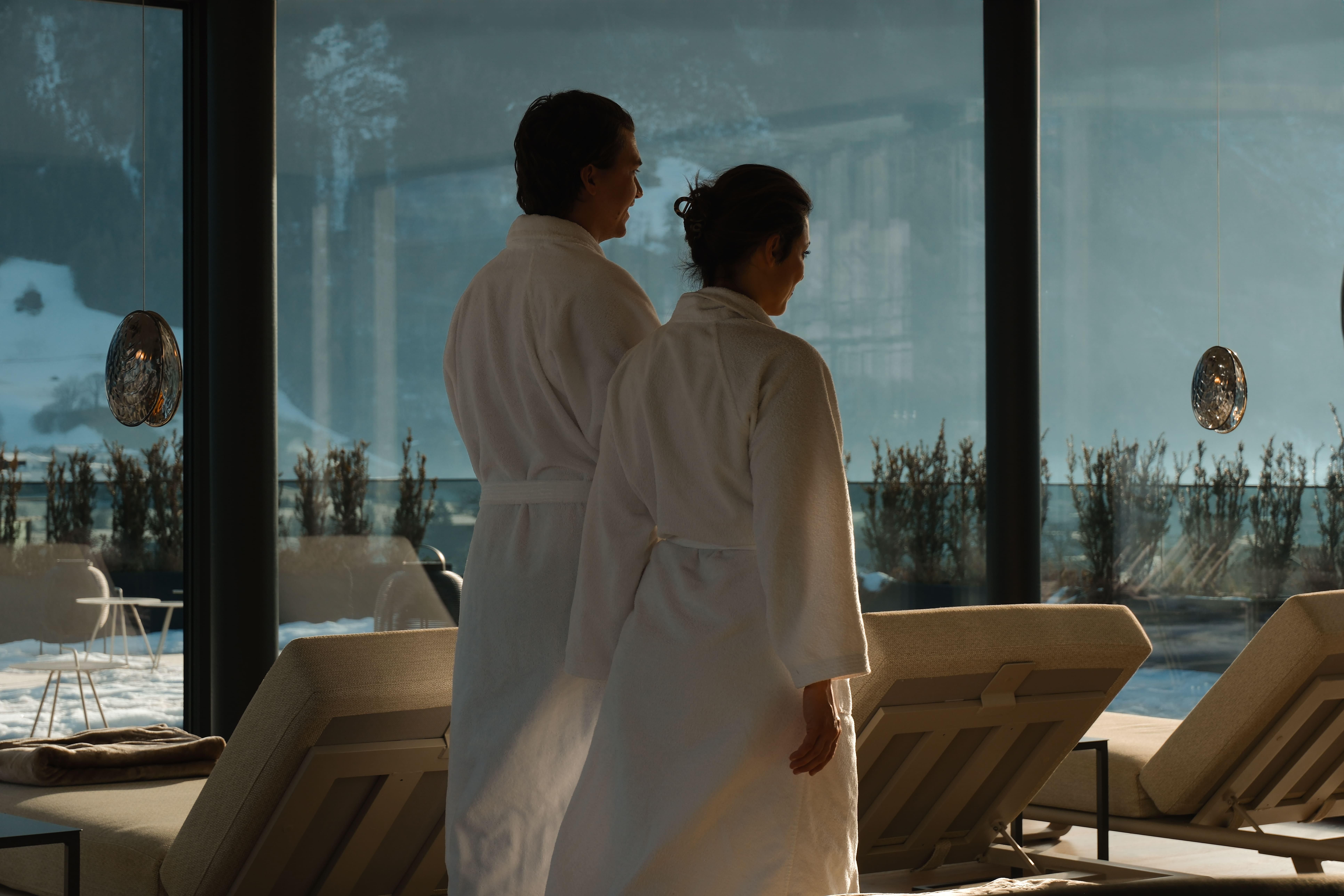 A couple in bathrobes stands in front of large windows and looks outside. The view shows snow and a relaxing atmosphere.