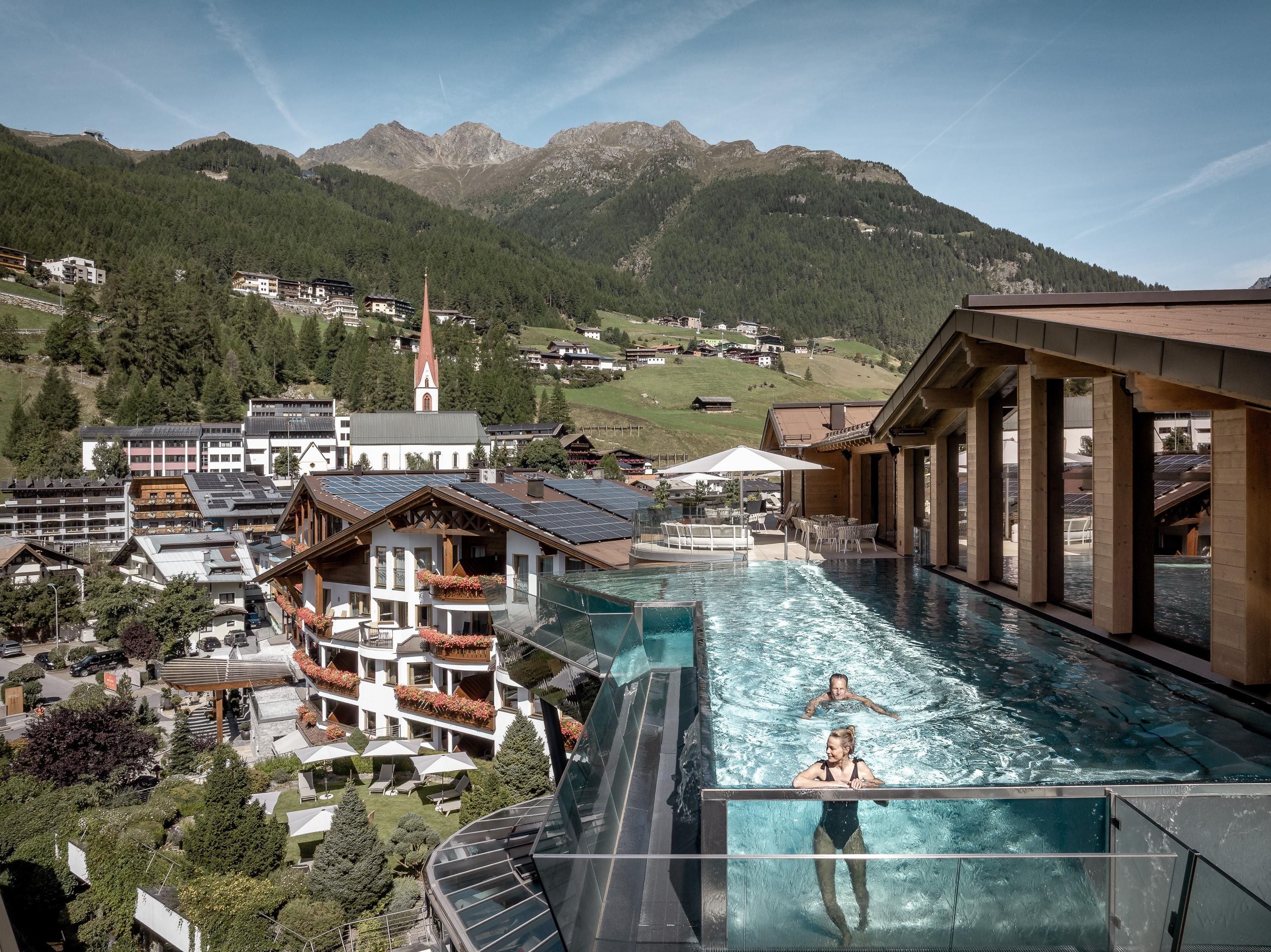 An infinity pool with a view of the mountains and the surrounding buildings. Two people are swimming in the water while the sun is shining.