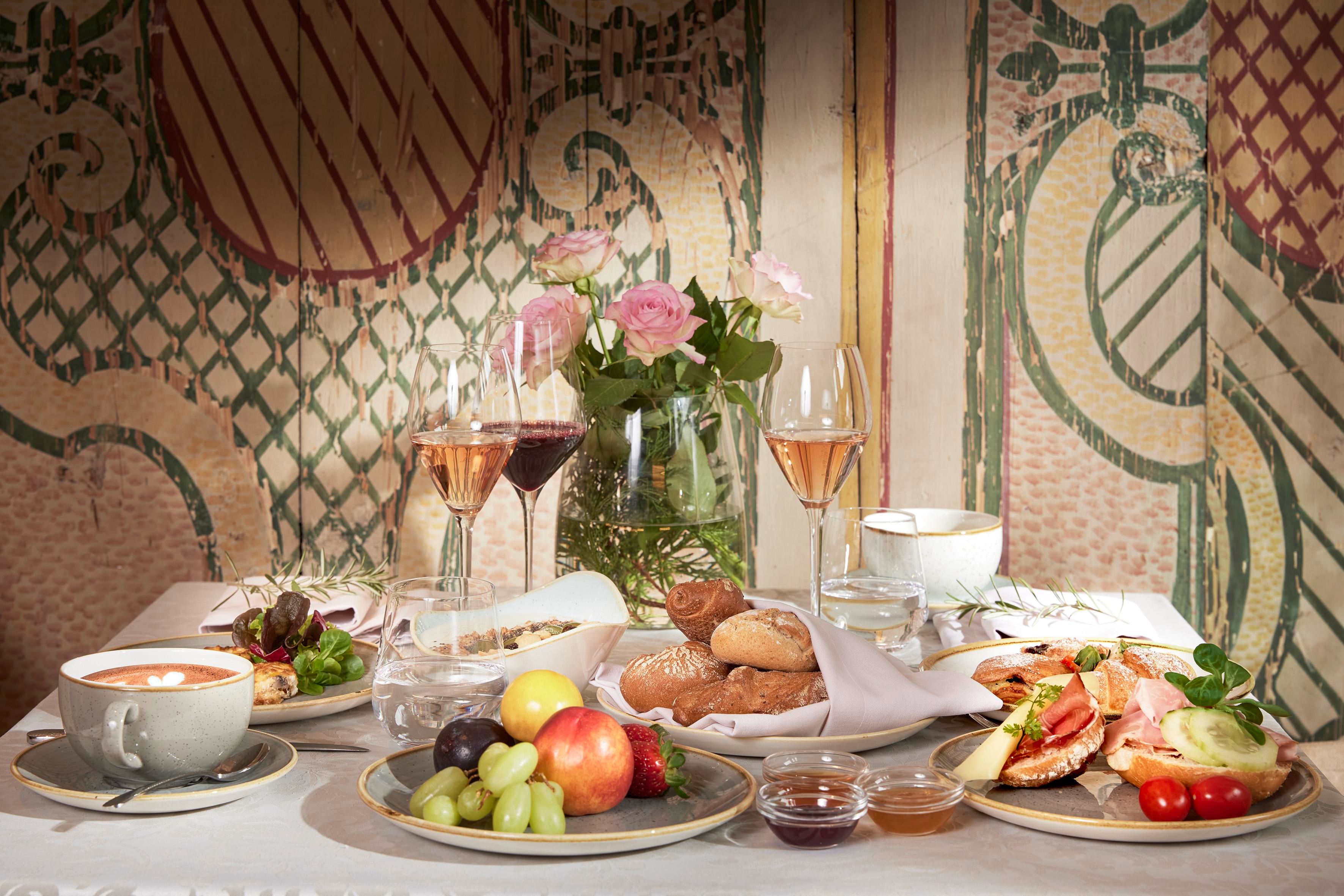A beautifully set table with various dishes, including fruit, pastries, and a soup. In the background, there are wine and a vase with flowers.