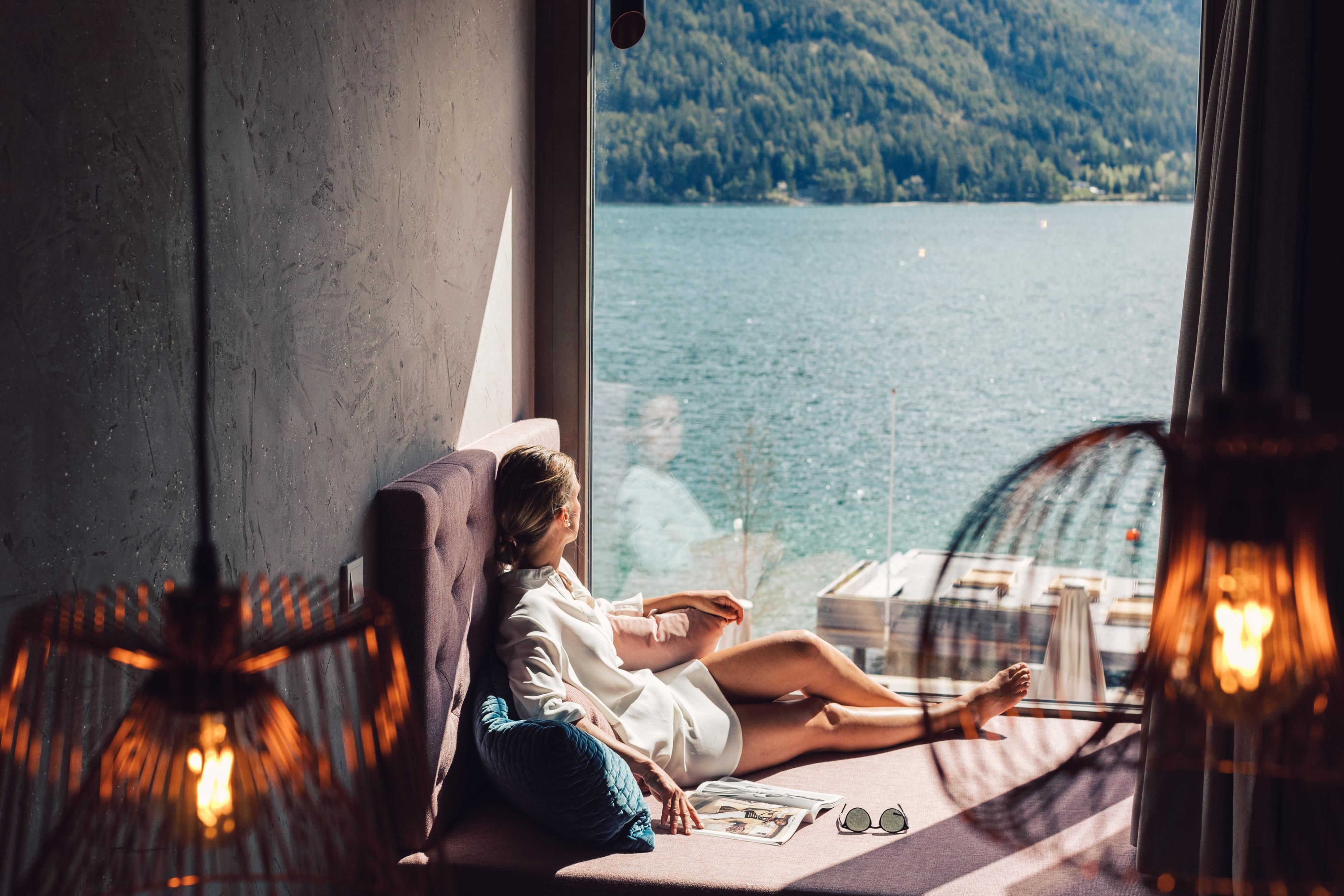 A woman relaxes by the window overlooking a lake. The surroundings are bright and modern, with warm light from lamps.