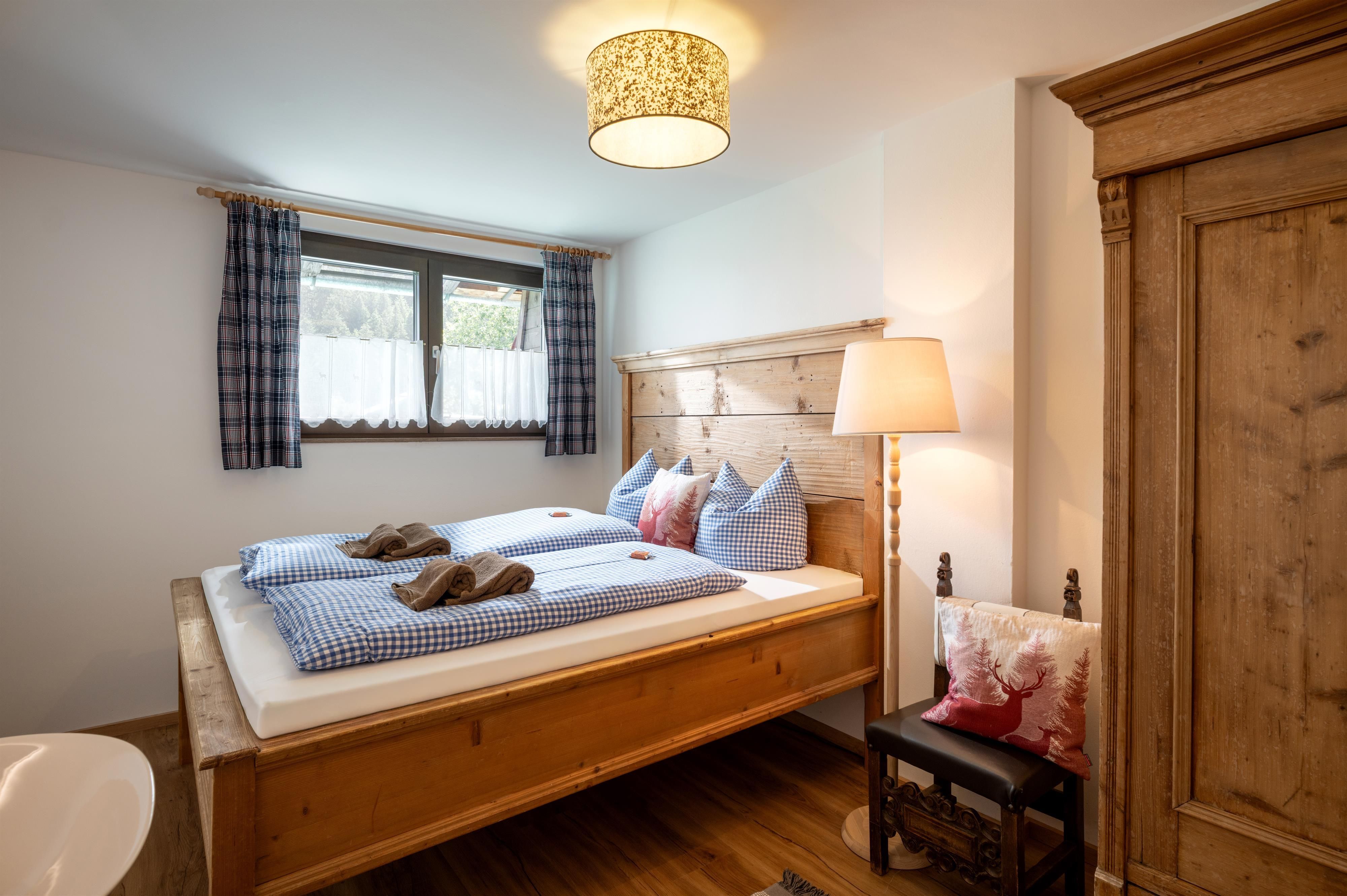 A cozy bedroom with a wooden bed and blue cushions. The room has a window with curtains and warm lighting.