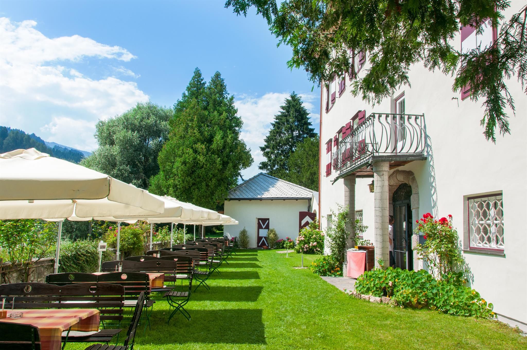 A charming garden view with umbrellas and wooden tables. In the background, an elegant building and green trees can be seen.