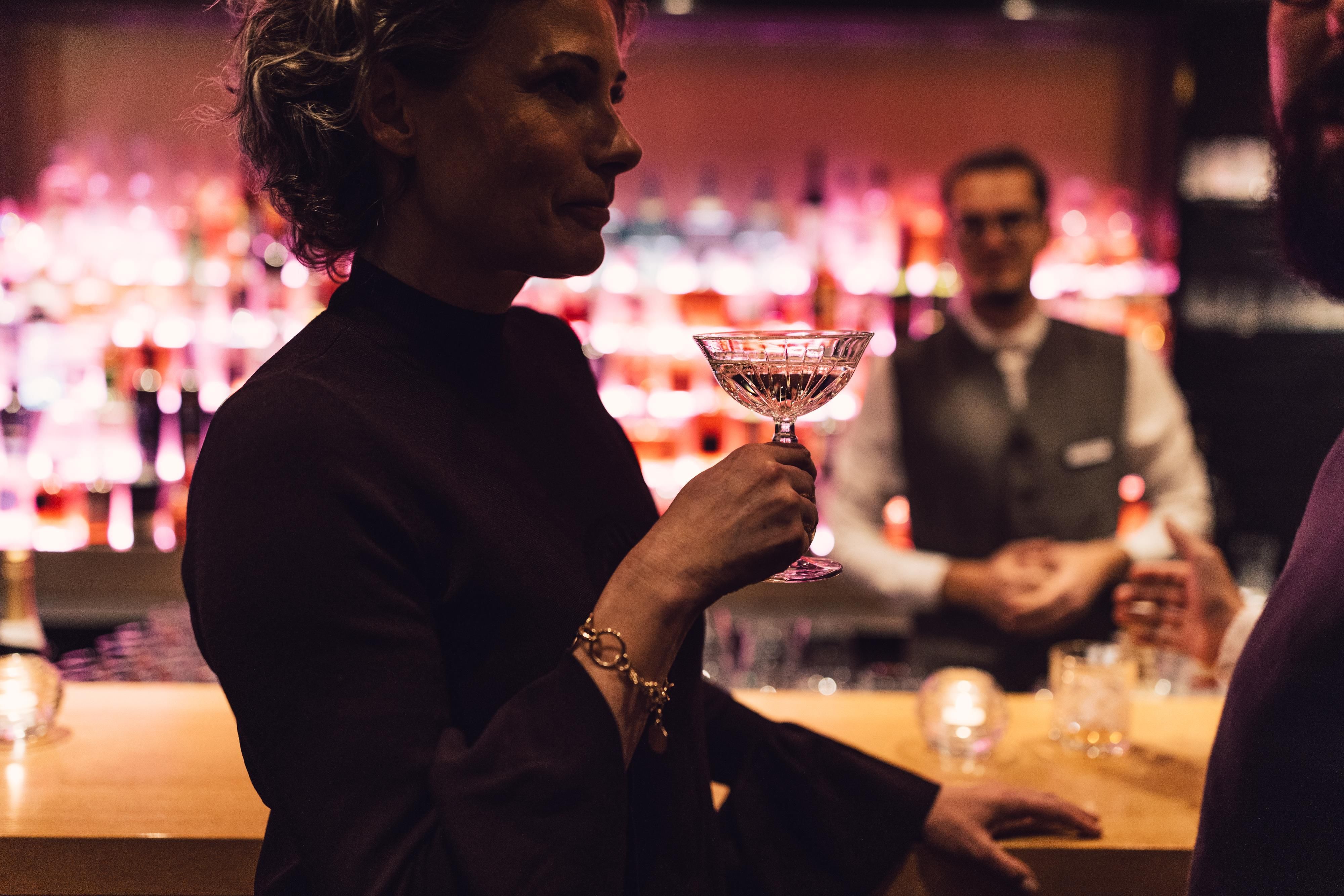 An elegantly dressed woman holds a cocktail glass in a stylish bar. In the background, the bartender is visible, surrounded by colorful drinks.