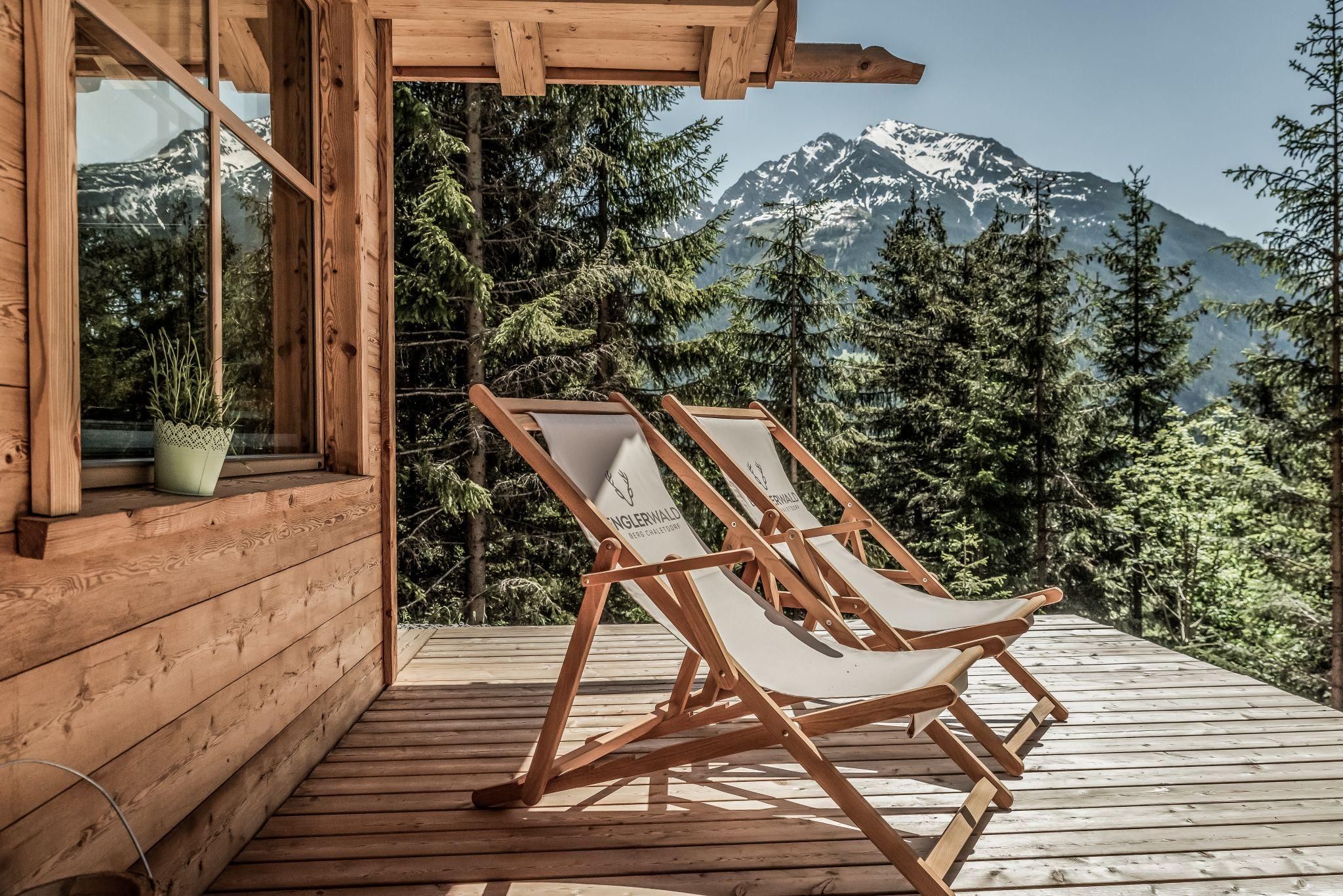 A cozy terrace with two loungers overlooks a wooded mountain. In the background, snow-capped peaks are visible.