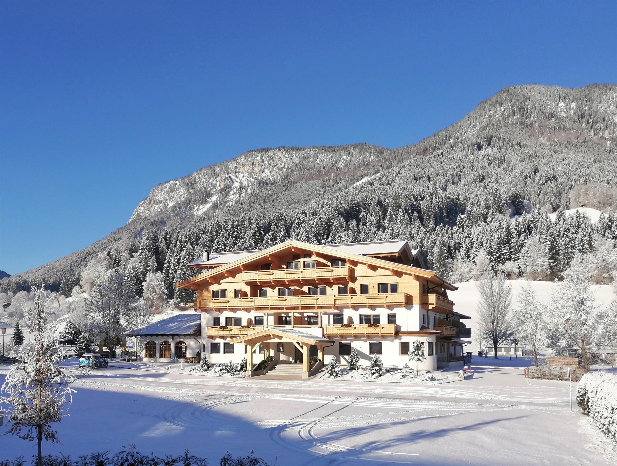 A charming hotel in winter, surrounded by snow-covered mountains and trees. The clear blue sky creates a soothing atmosphere.