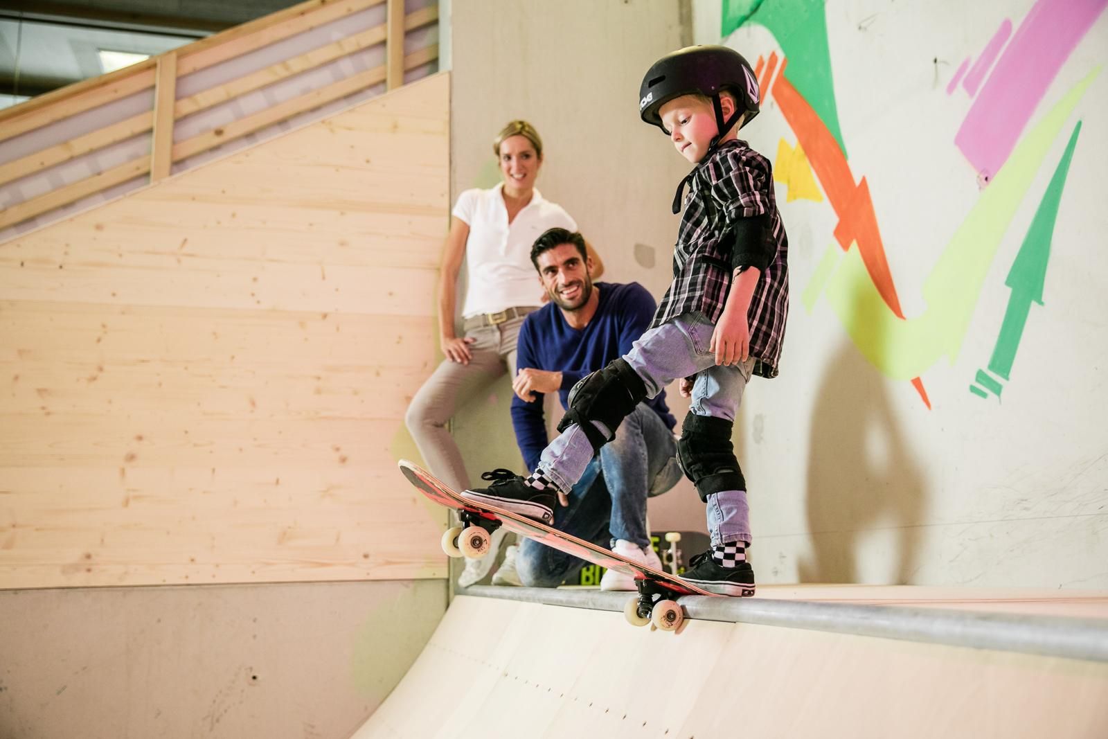 A boy is skateboarding on a ramp and wearing protective gear. In the background, there are two adults watching him.