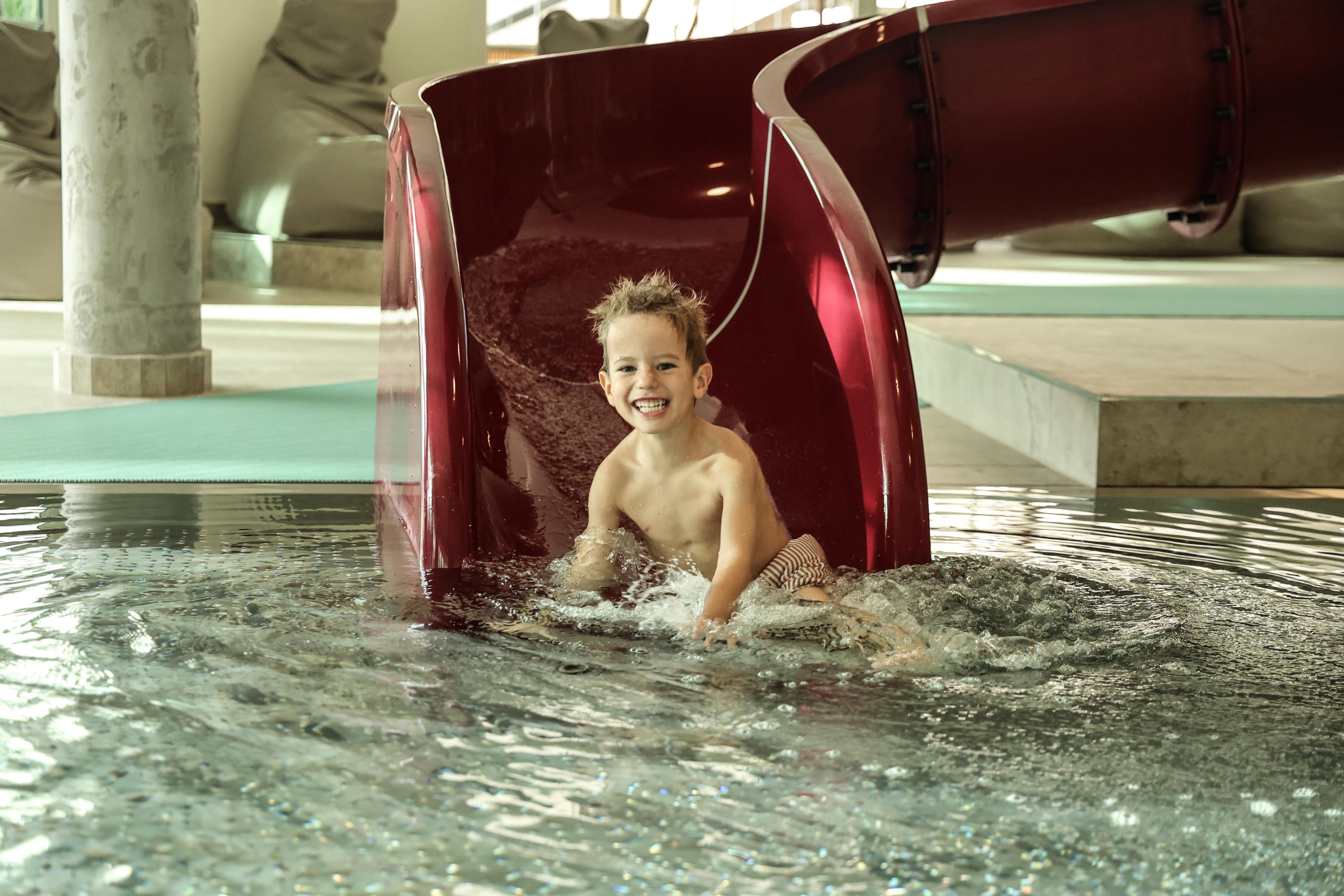 A cheerful boy sits in a pool at the end of a red slide. He is having a lot of fun in the water.