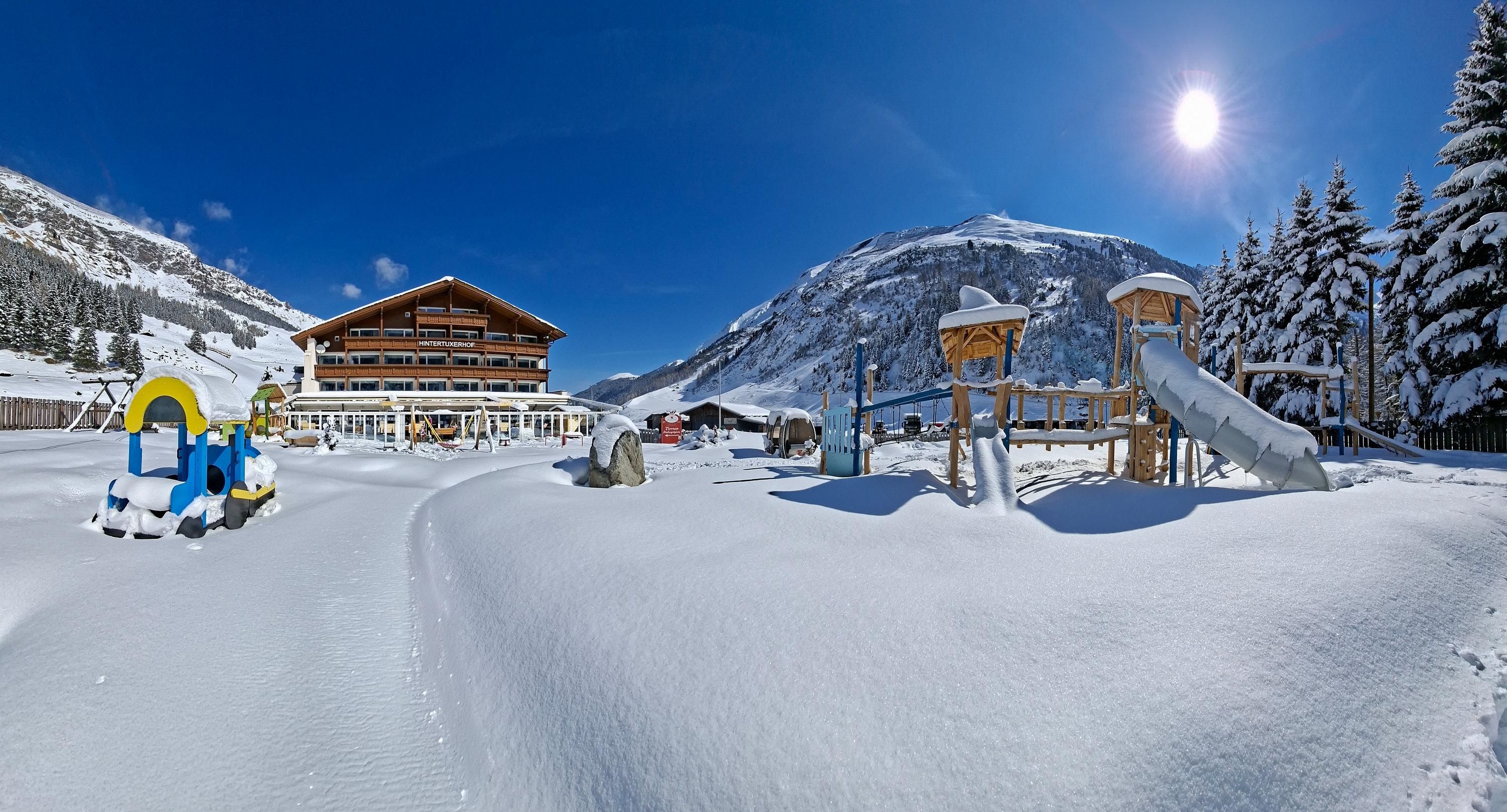 A snowy winter landscape with a playground and a building in the background. The sky is clear and the sun is shining.