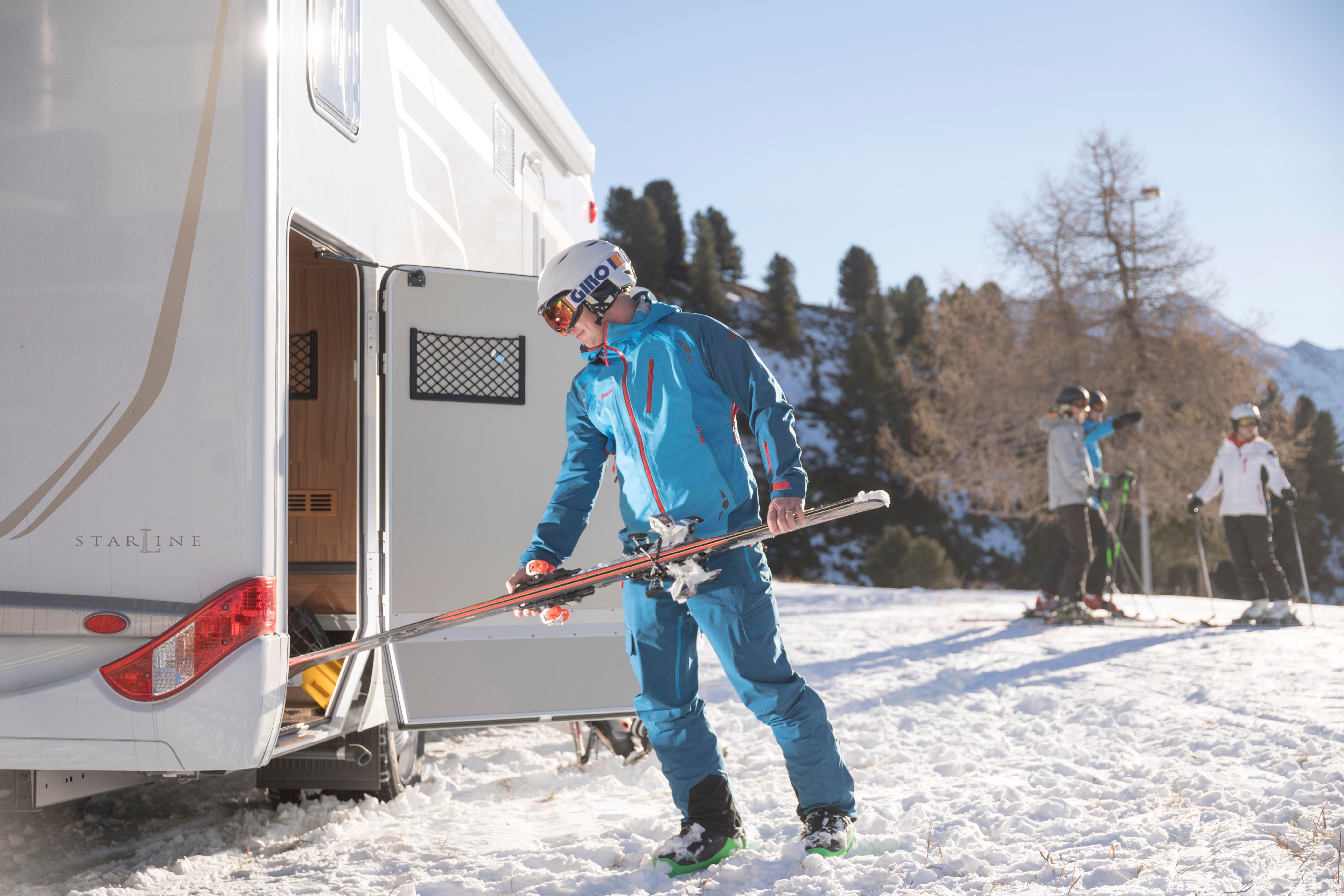 A skier in blue gear is preparing to take his skis out of a camper van. In the background, other skiers can be seen on a snow-covered slope.