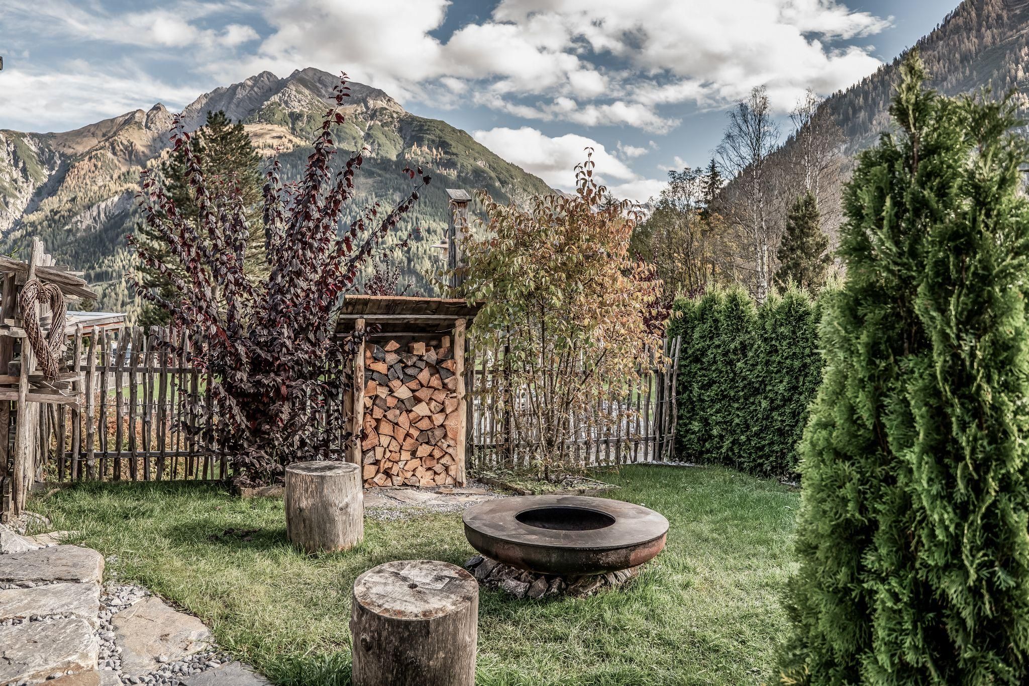 An idyllic garden with a grill bowl, surrounded by trees and a woodpile. In the background, majestic mountains rise under a cloudy sky.