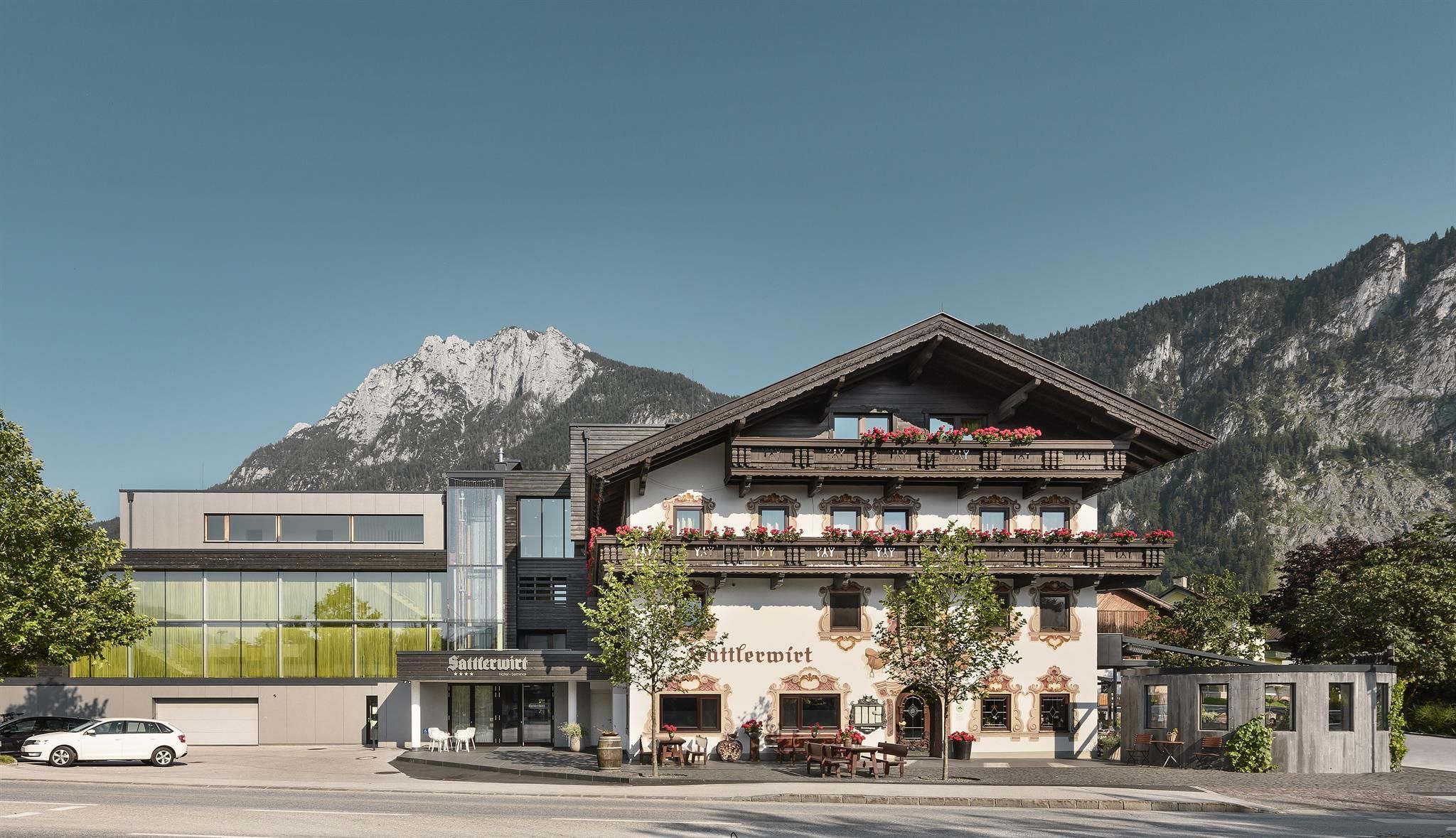 A modern hotel building with a traditional alpine style. In the background, majestic mountains rise under a clear sky.