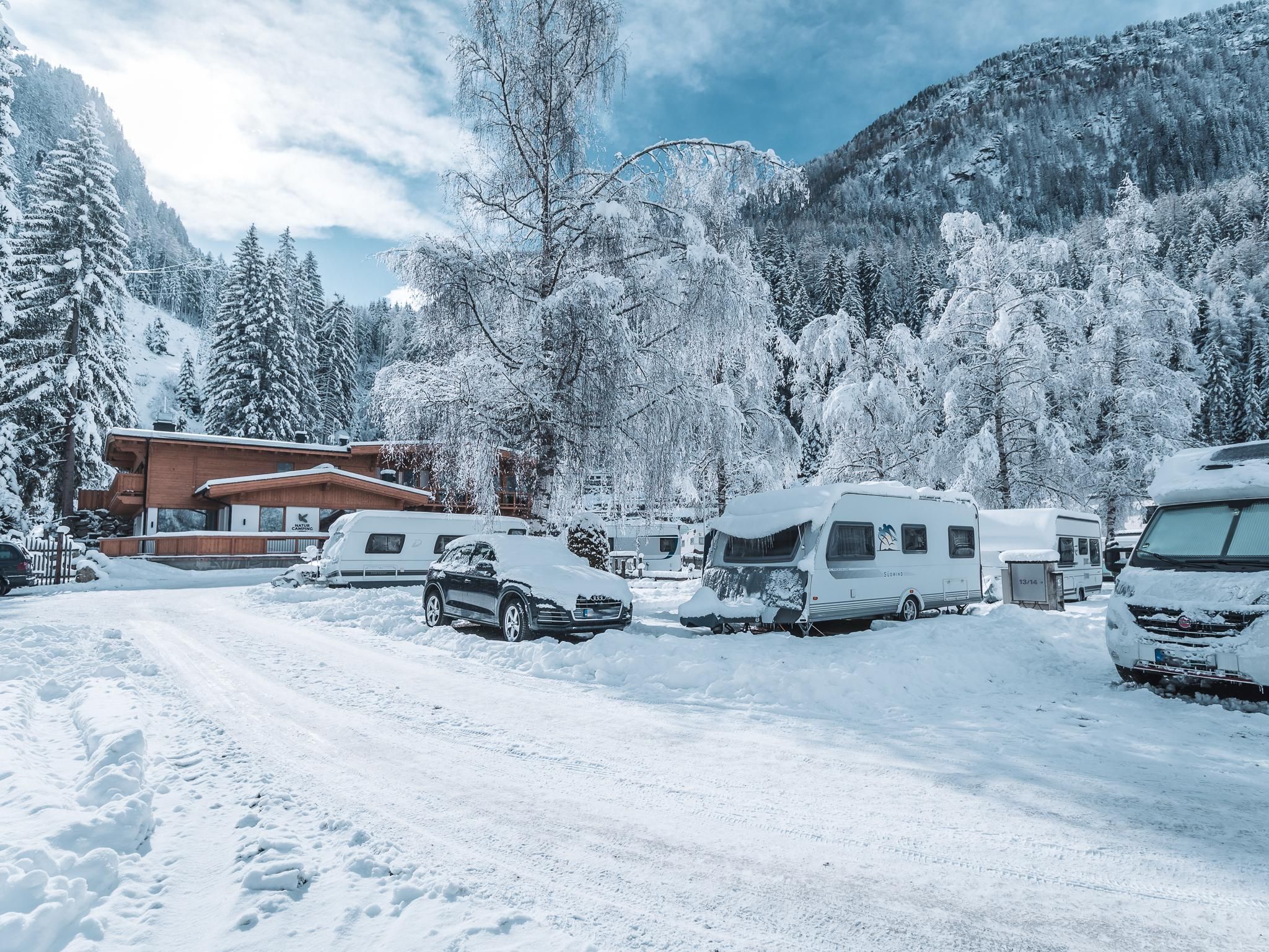 A snowy campsite with caravans and parked cars. In the background, there are snow-covered mountains and trees.