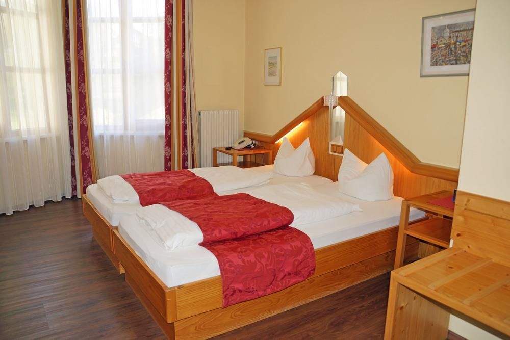 A cozy hotel room with two separate beds and warm wooden details. Bright windows allow plenty of daylight in.
