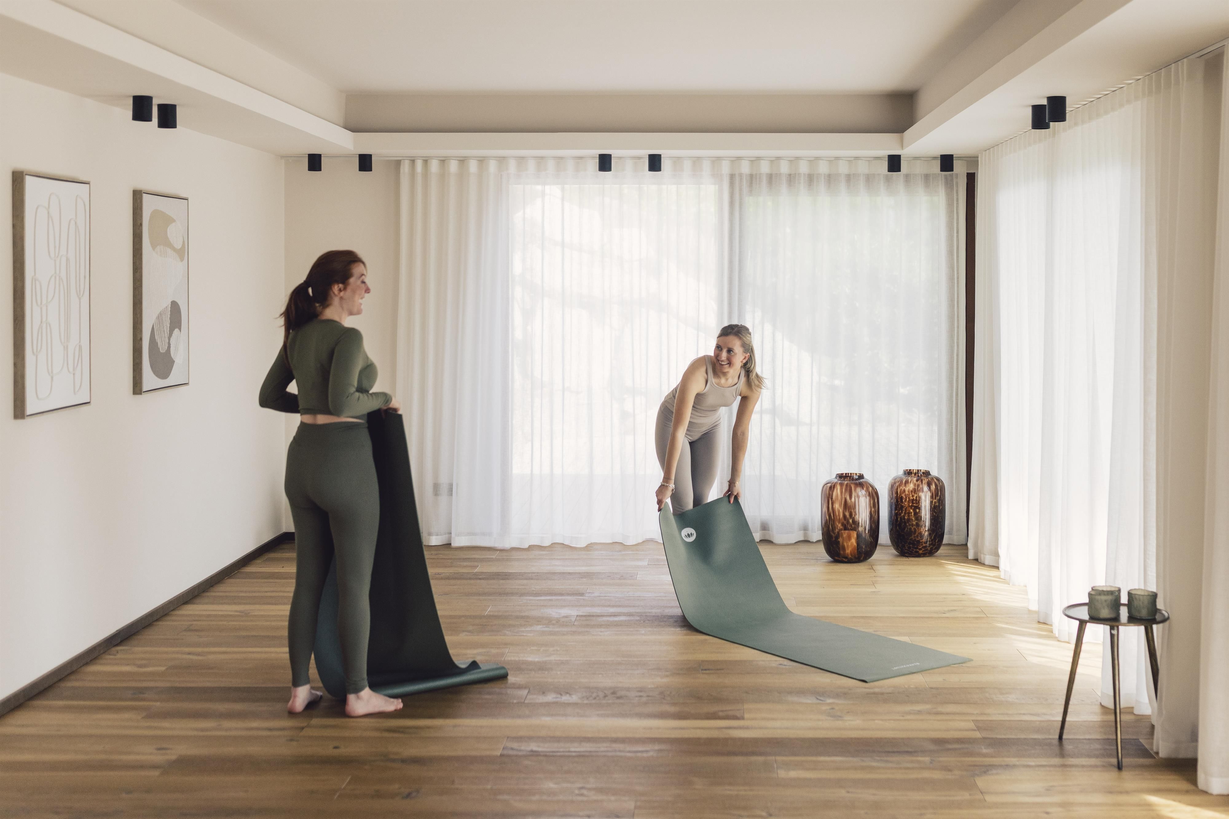 Two women are preparing for a yoga session in a bright room with a wooden floor. They are unrolling their yoga mats as the room is filled with natural light.