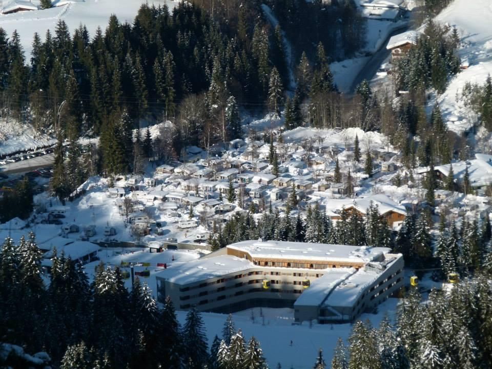 A snowy landscape with a modern hotel and a settlement of holiday homes. Surrounded by dense, green forests and mountains.