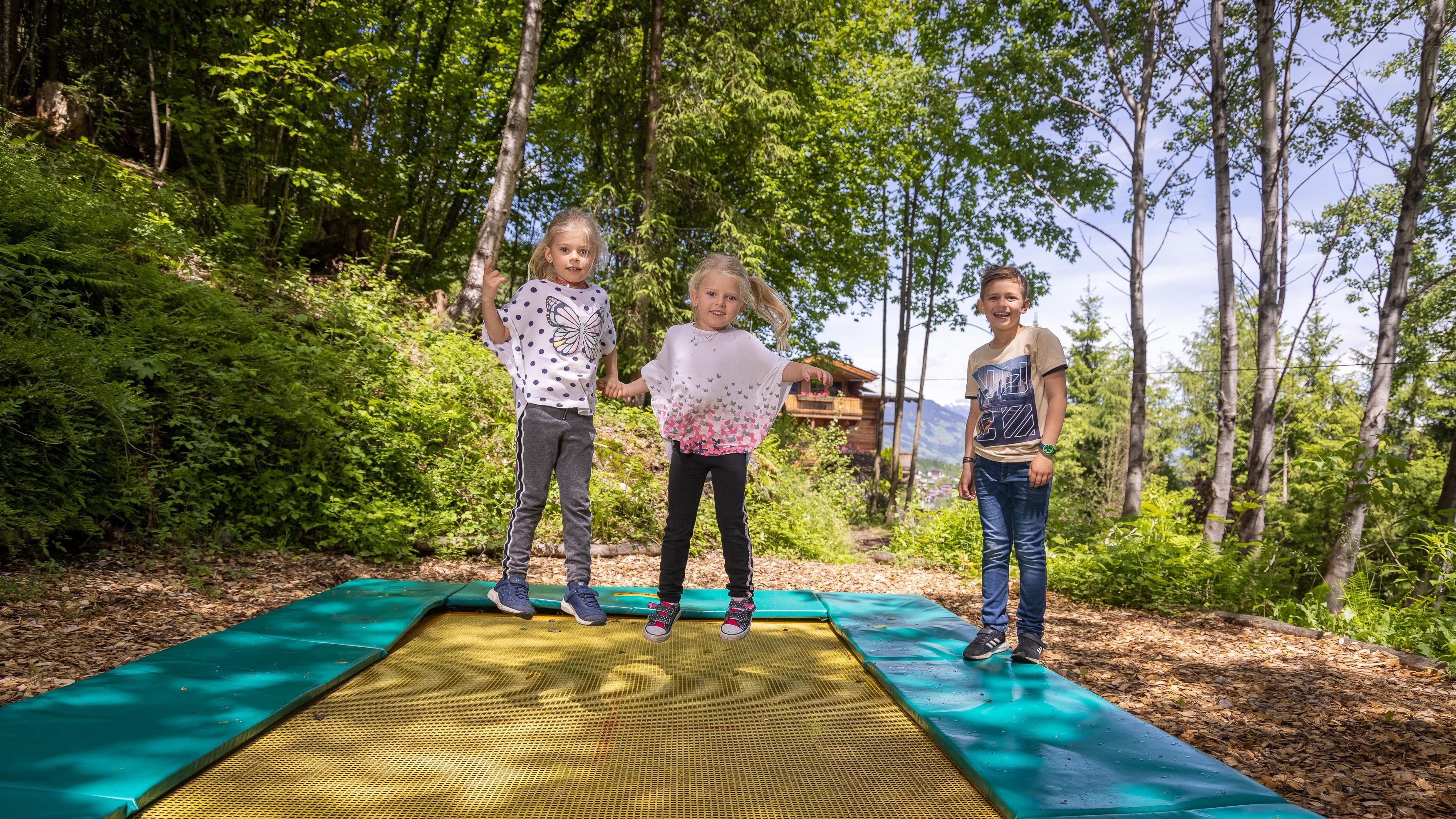 Three children are standing on a trampoline outdoors. Around them are trees and green foliage.