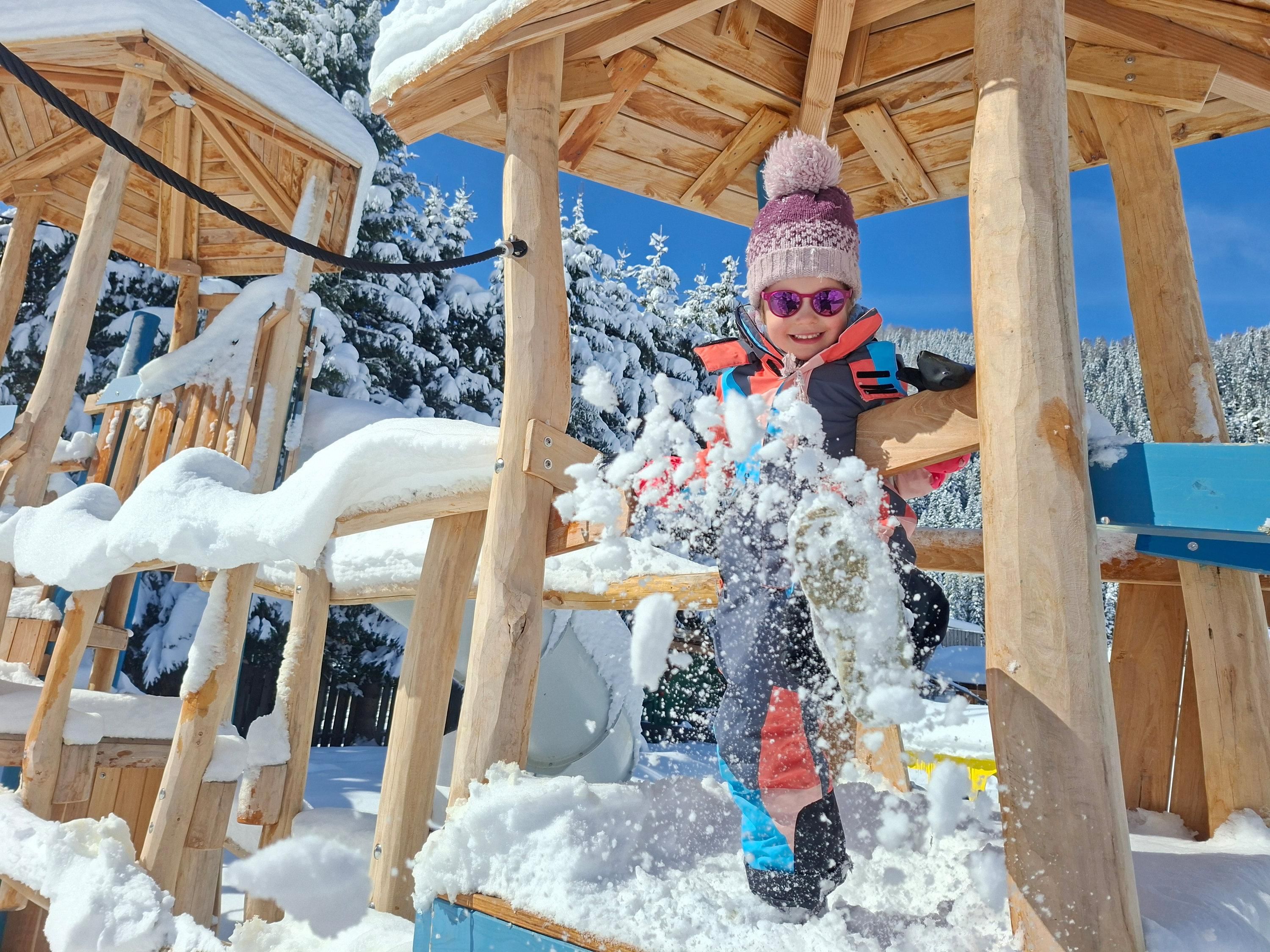 A child is happily playing in the snow at a playground. The surroundings are wintry with plenty of snow and a blue sky.