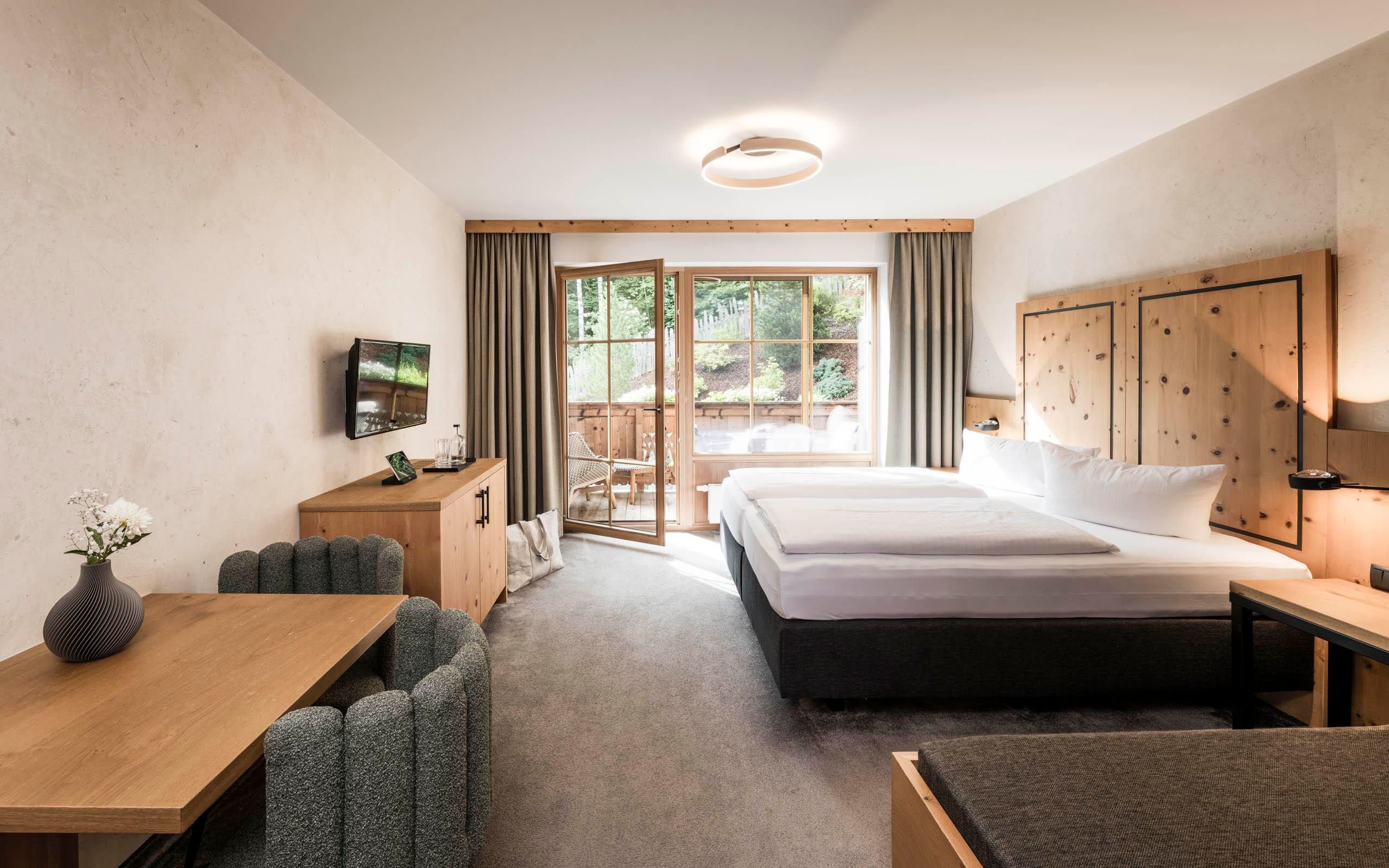 A modern hotel room with a large bed and wooden furniture. Large windows offer a view of the balcony and the garden.
