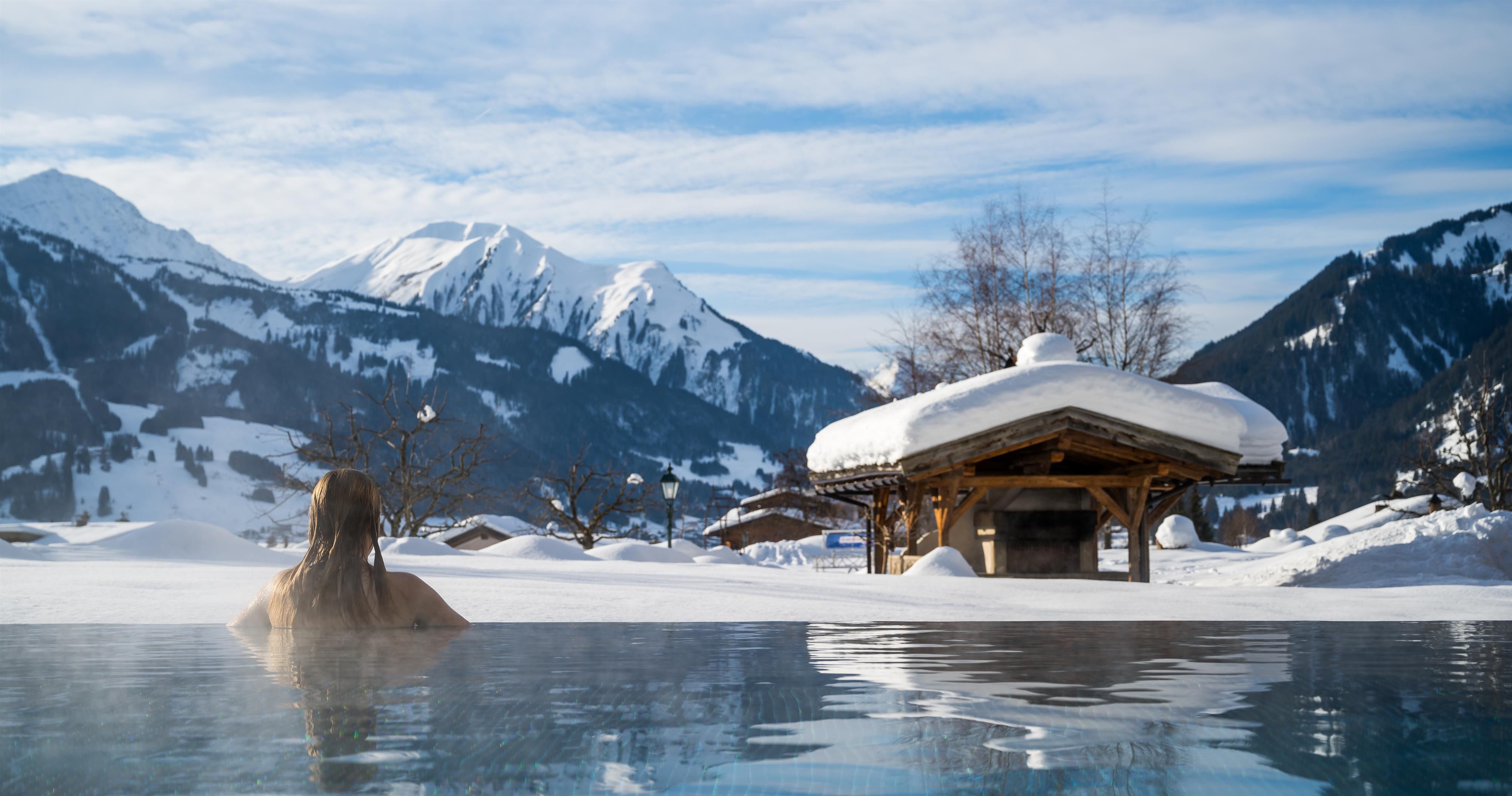 A relaxing thermal pool in a snow-covered landscape. In the background, majestic mountains and a cozy log cabin can be seen.