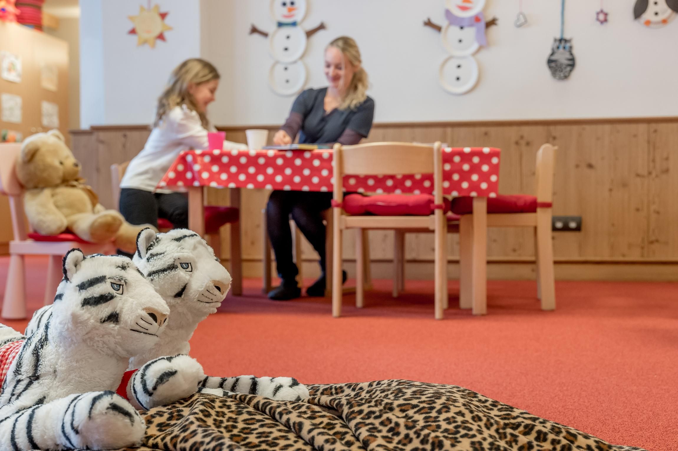 A cozy playroom with a table and chairs. A girl and a woman are playing together while toy animals lie in the foreground.
