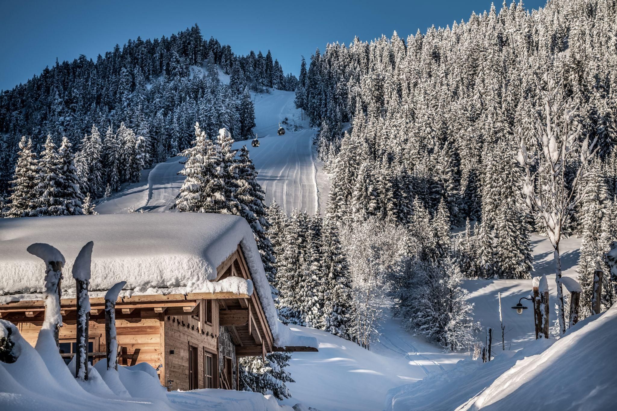 A picturesque winter landscape with snow-covered mountains and fir trees. In the foreground stands a cozy chalet.