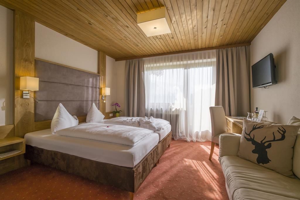 A cozy hotel room with a large bed and wooden ceiling. In front of the window is a bright curtain, and there is a seating area.