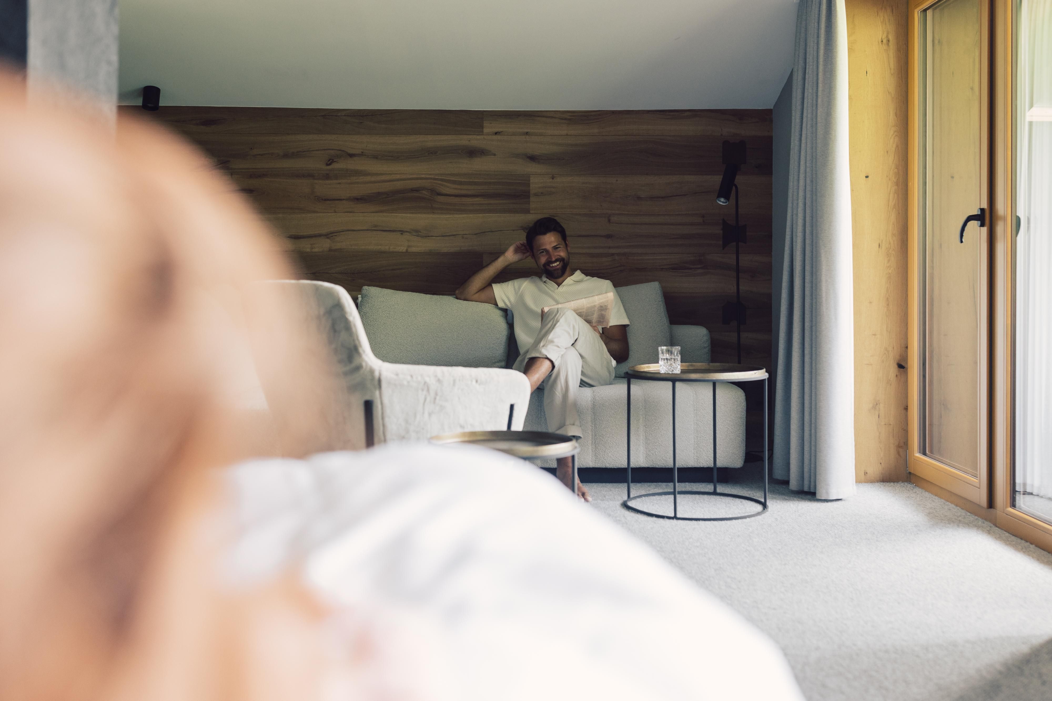 A man sits relaxed on a sofa in a modern room. Around him are large windows and wooden details that create a cozy atmosphere.