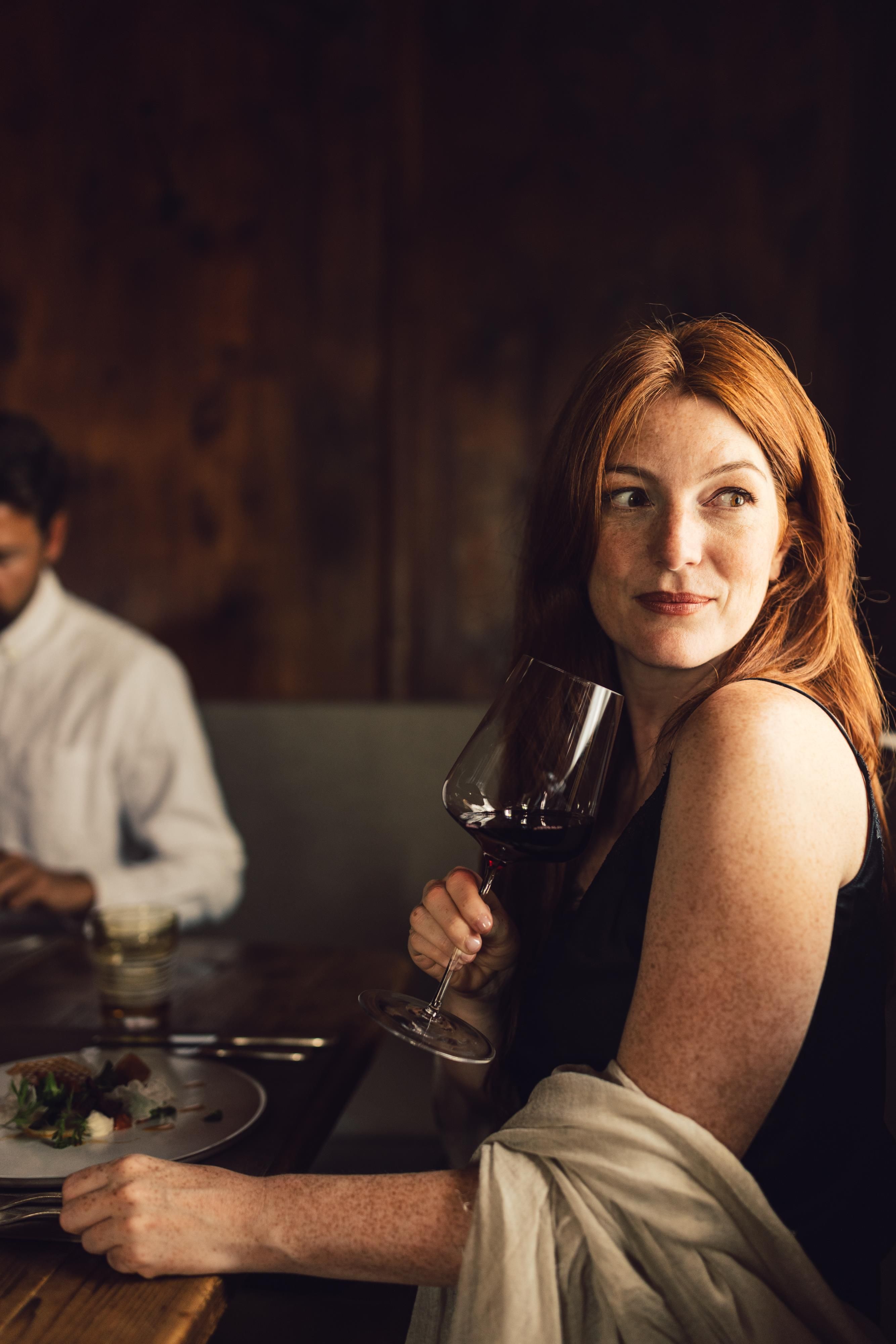 A woman with long, red hair holds a wine glass and looks pensively. In the background, a man can be seen sitting at the table and eating.