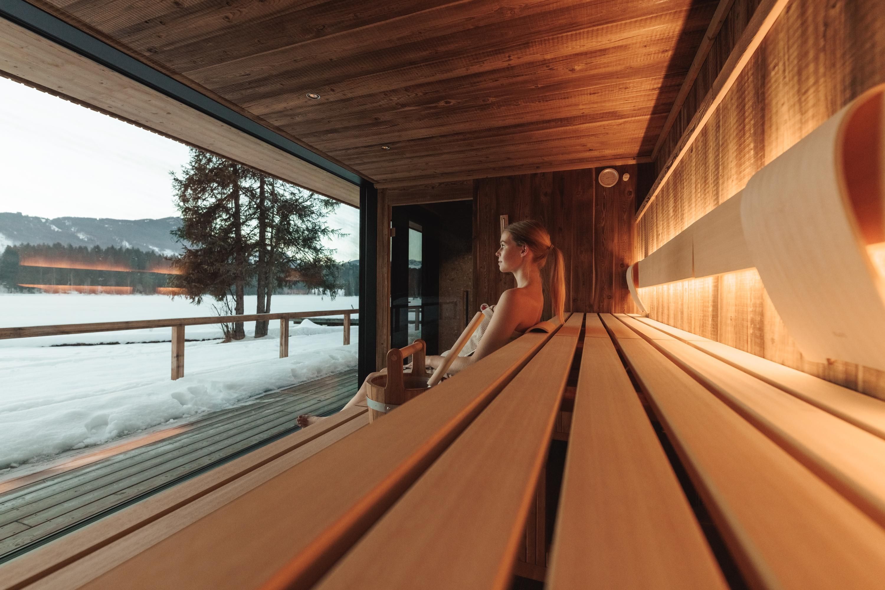 A modern sauna interior with wooden shelves and large windows. In the background, there is a winter landscape with snow and trees.