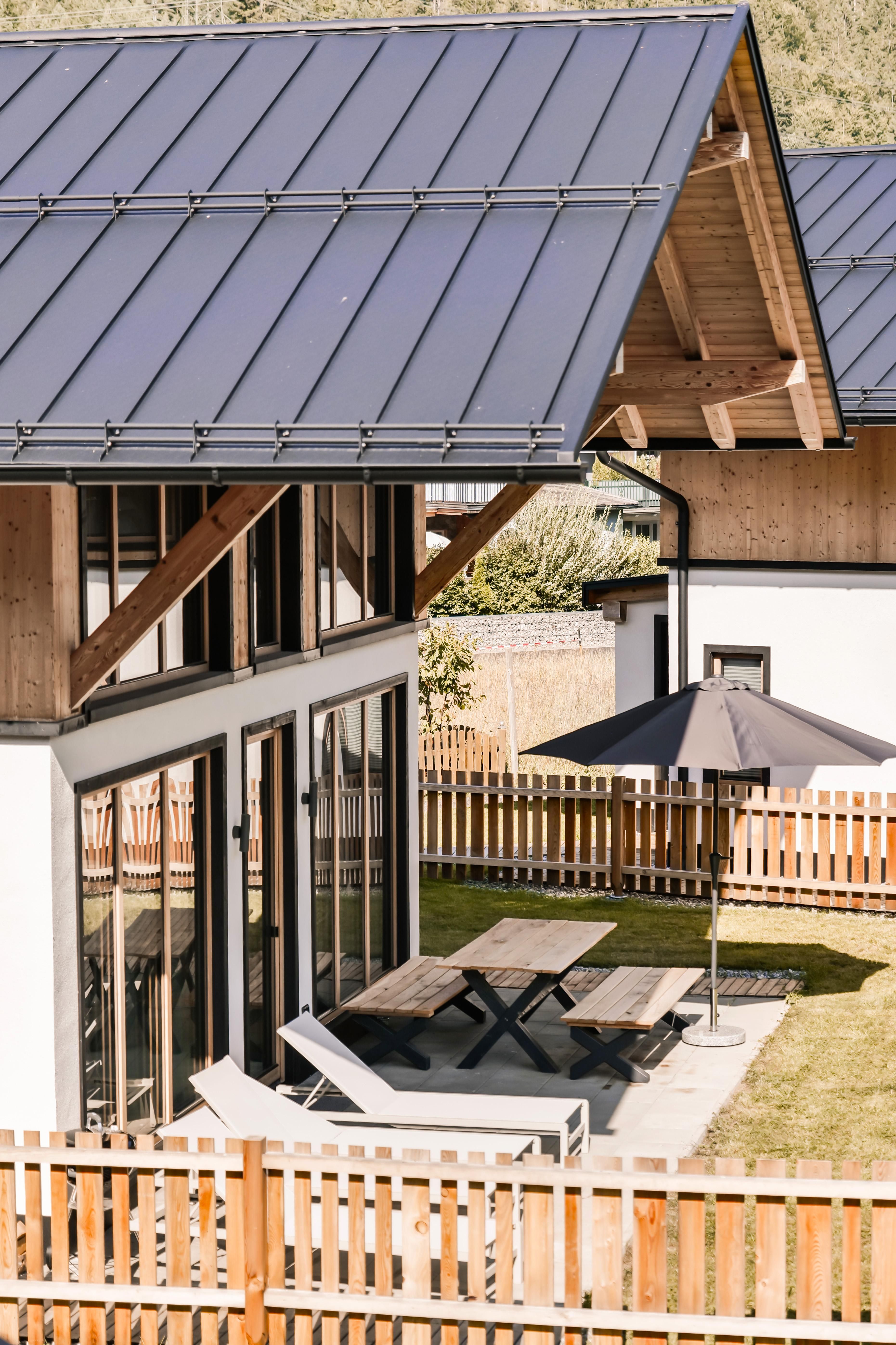 A modern wooden house with a sloped roof. Outside, there is a table with chairs and a sunshade in a well-maintained garden.