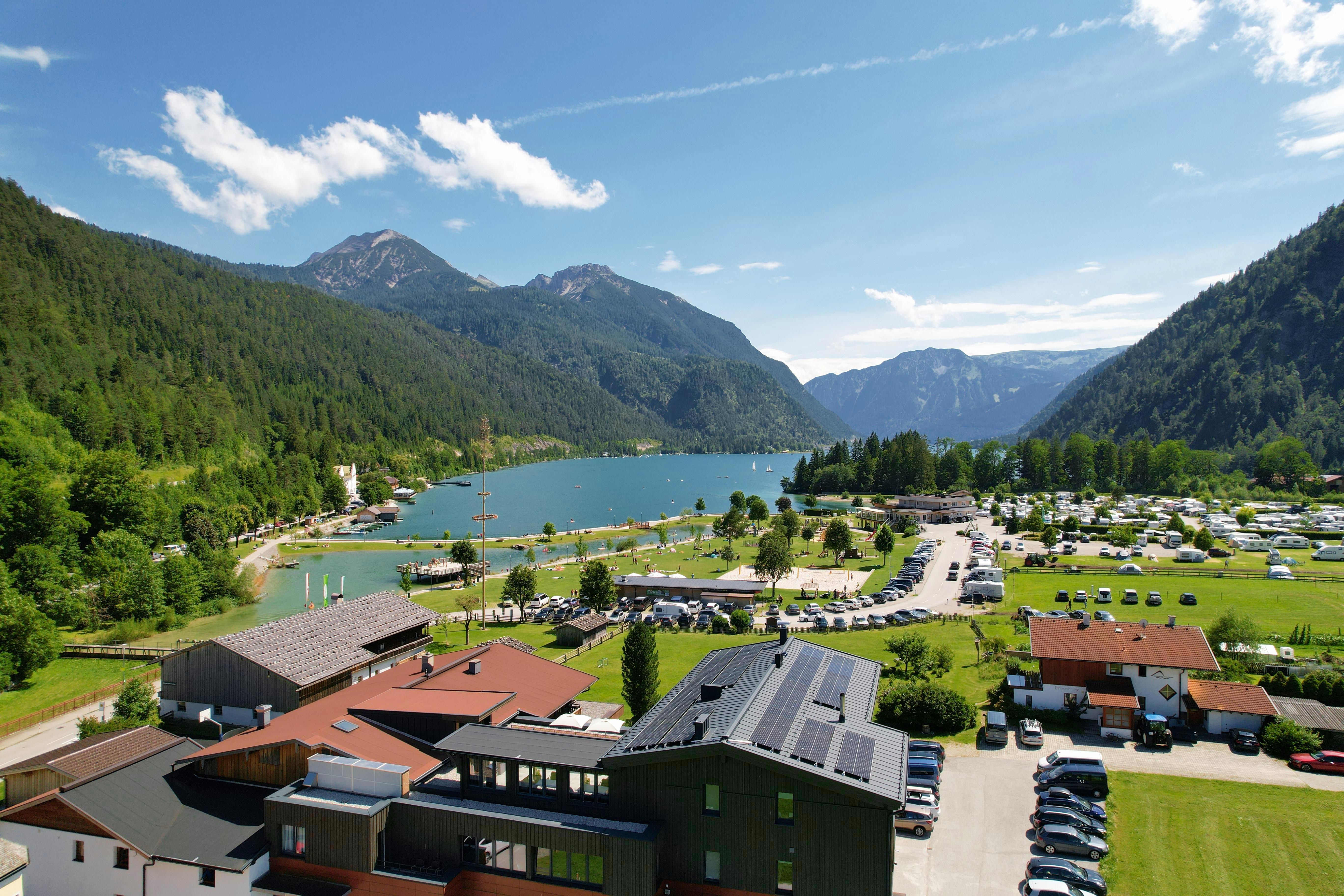 A picturesque lake surrounded by mountains and green forests. In the foreground, there are beautiful buildings and a campsite.