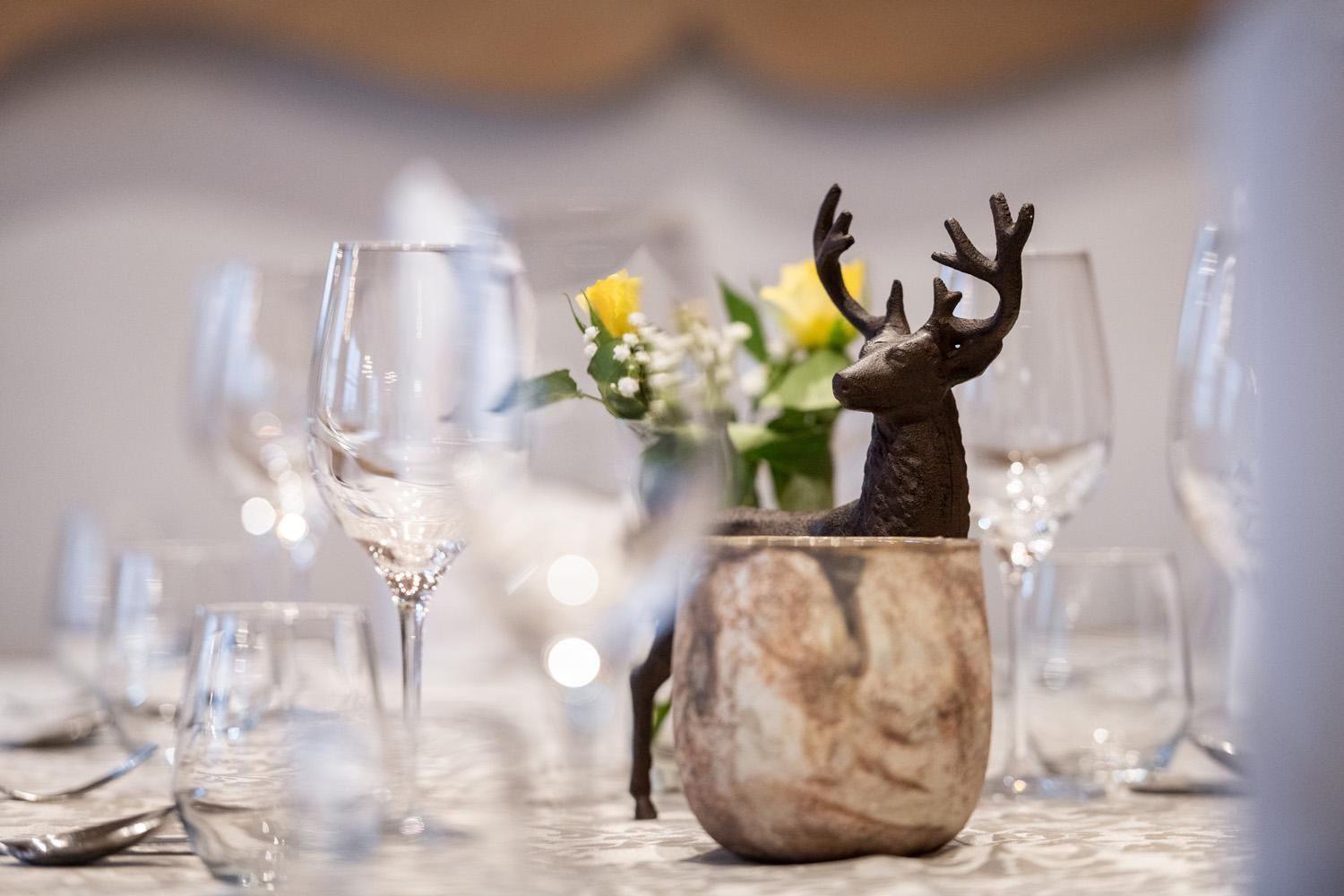 An elegant table with wine glasses and a decorative deer figure in a stone container. In the background, yellow flowers can be seen, adding a touch of brightness to the ambiance.