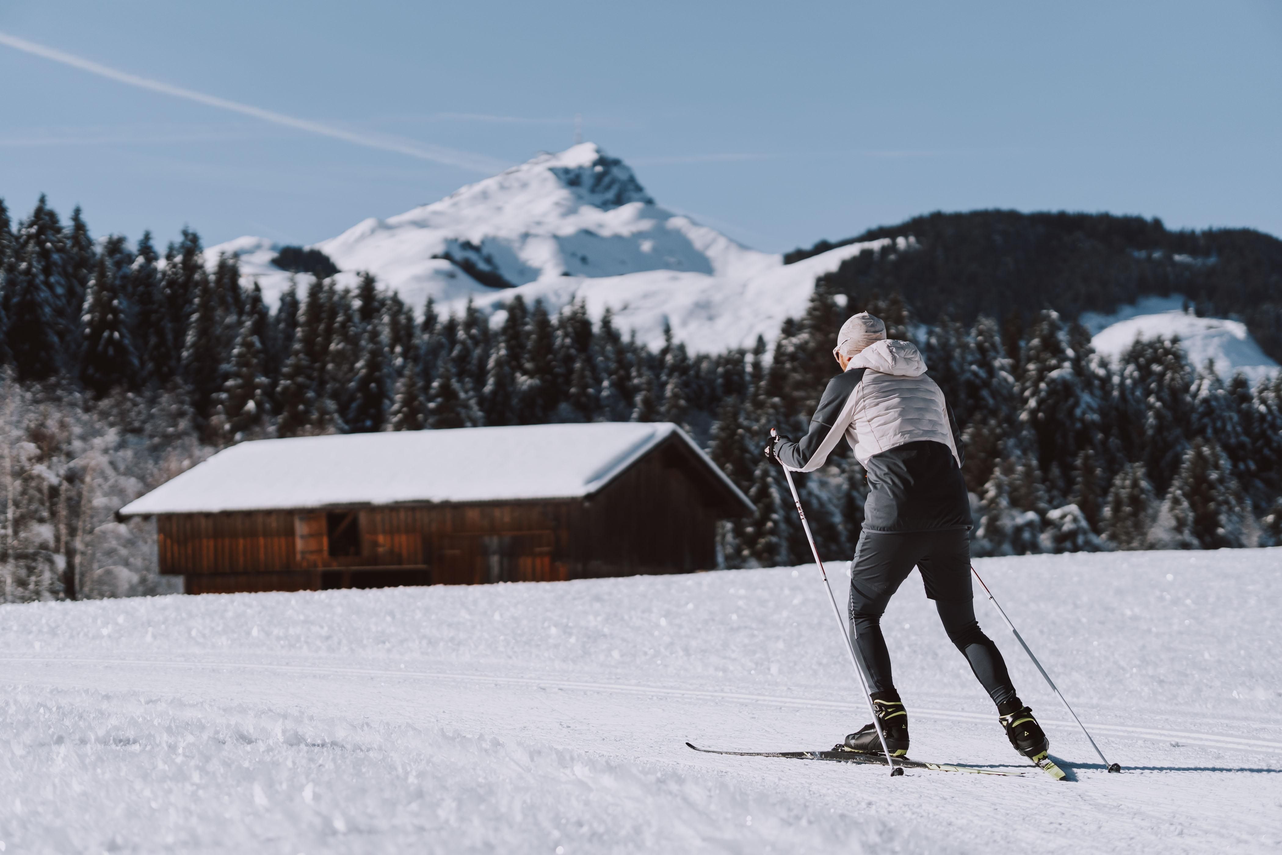 A skier glides through the snowy landscape. In the background, snow-covered mountains and a wooden barn are visible.