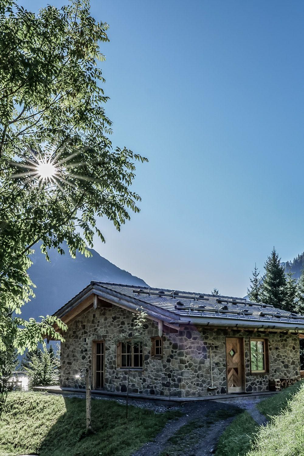 Stone chalet surrounded by trees and green meadow. The sun is shining on the roof and the surrounding landscape.