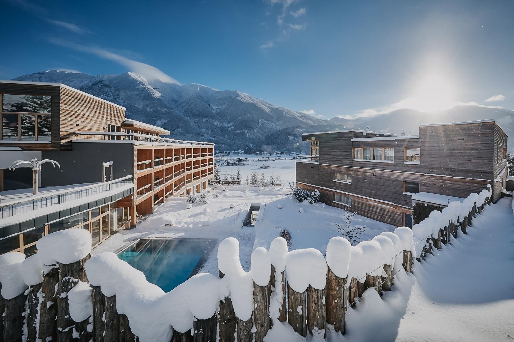 A snowy winter landscape with modern wooden buildings and a pool. In the background, mountains and a clear sky are visible.
