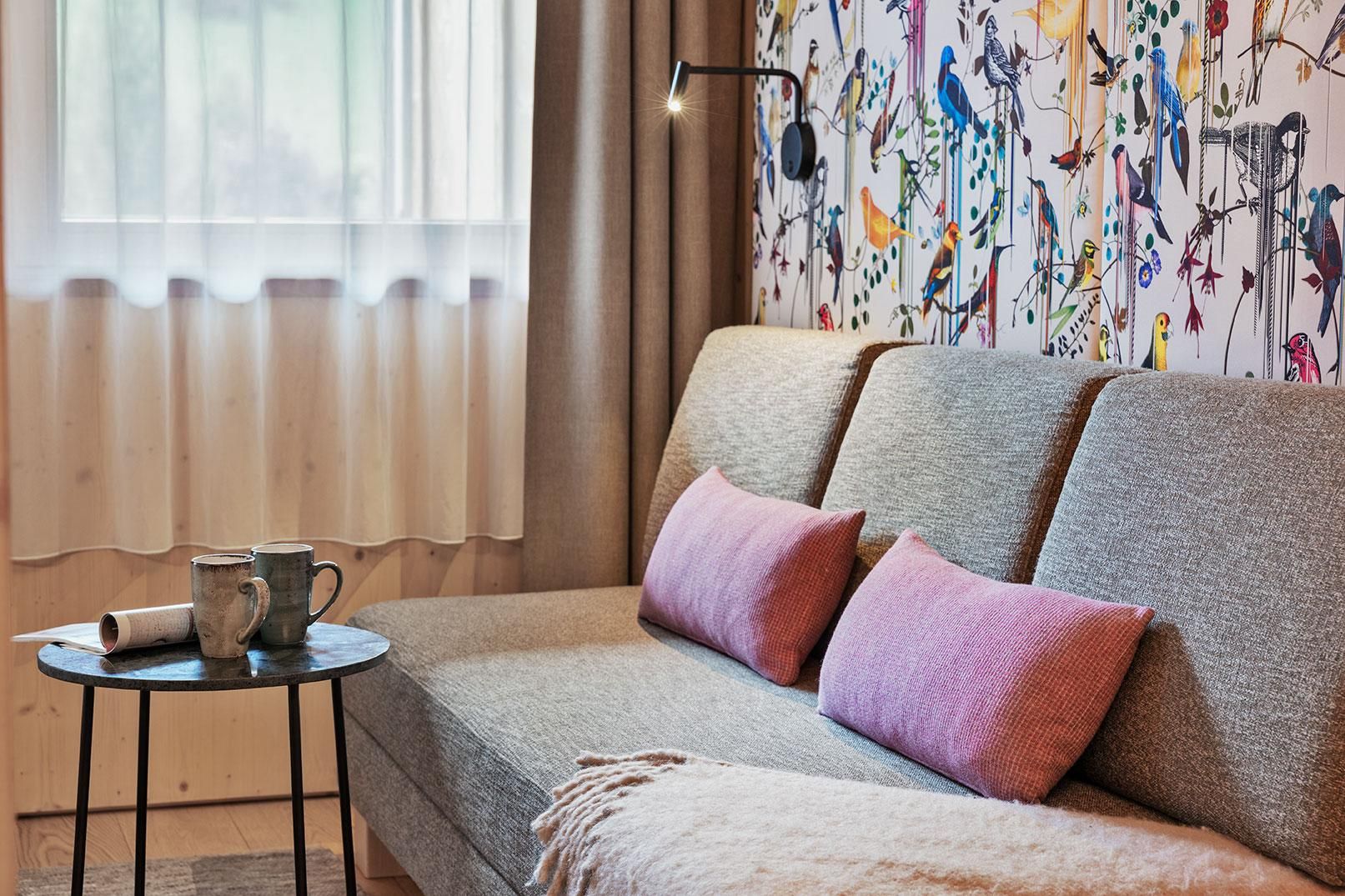 A cozy living room with a gray sofa and pink cushions. In the background, there is a colorful bird wall design and a small table with cups next to it.