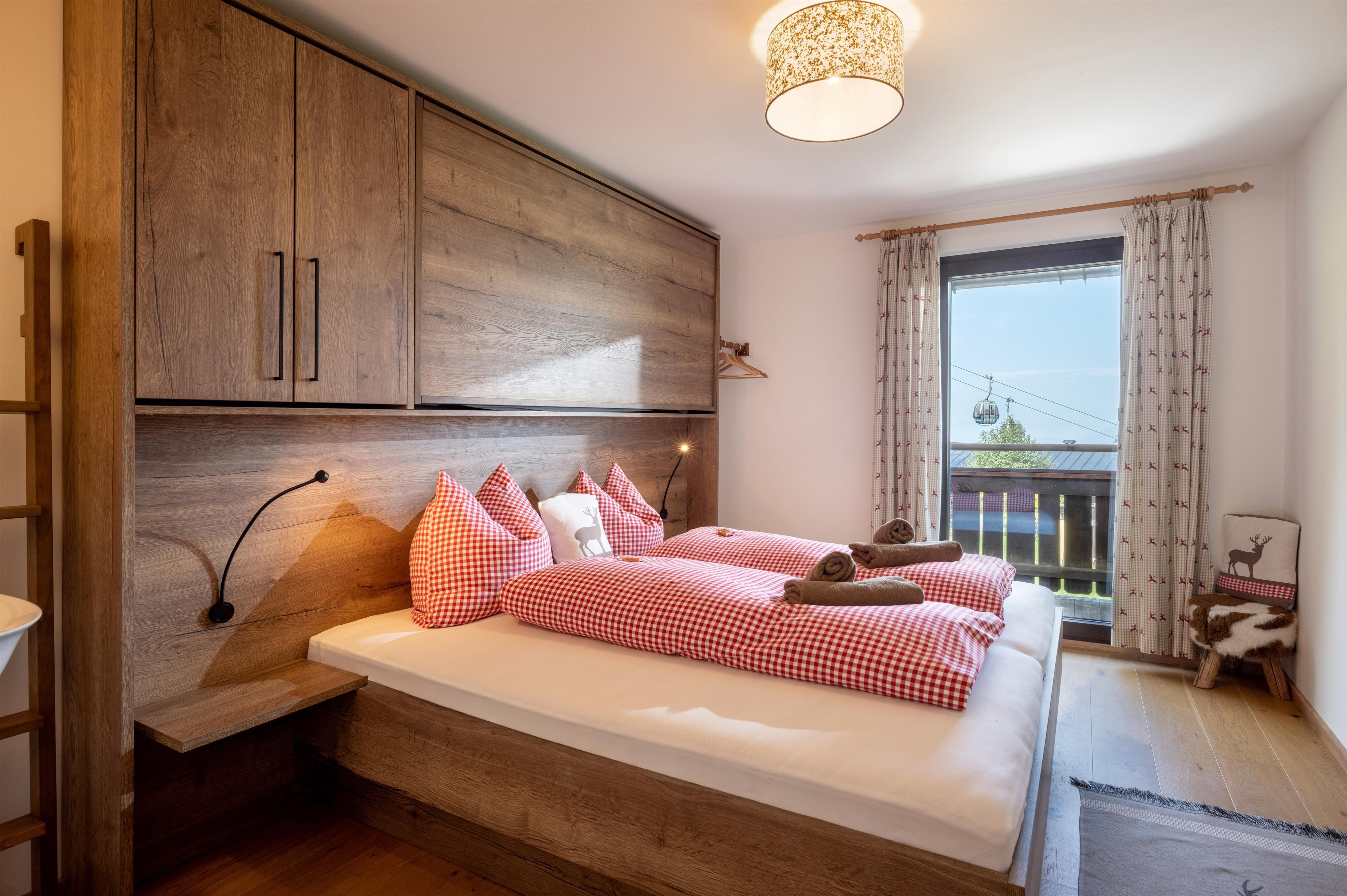 A cozy bedroom with a double bed and red-white cushions. The room has large windows and modern wooden furniture that create an inviting atmosphere.