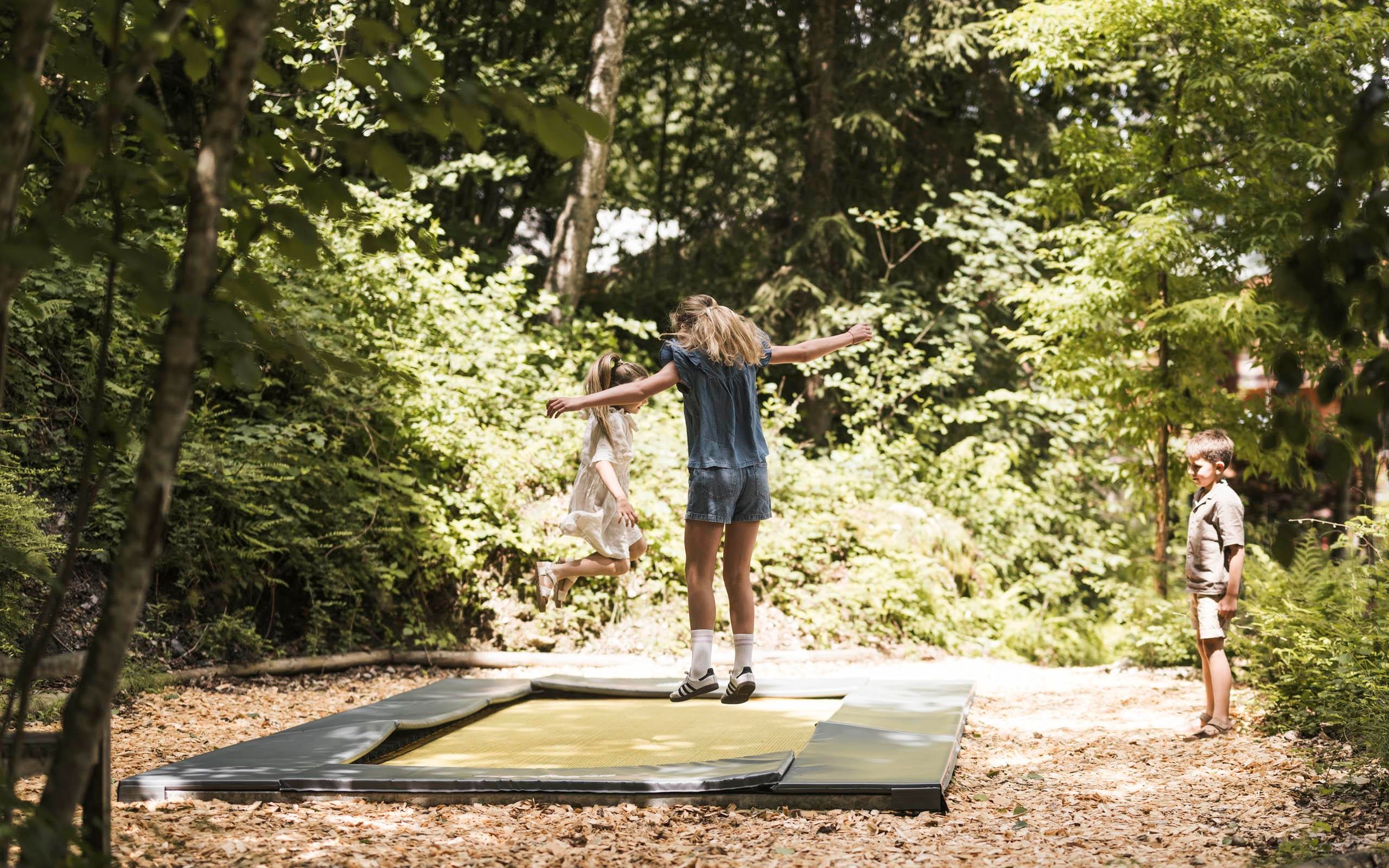 A girl is happily jumping on a trampoline while a boy watches beside her. The surroundings are filled with trees and leaves.