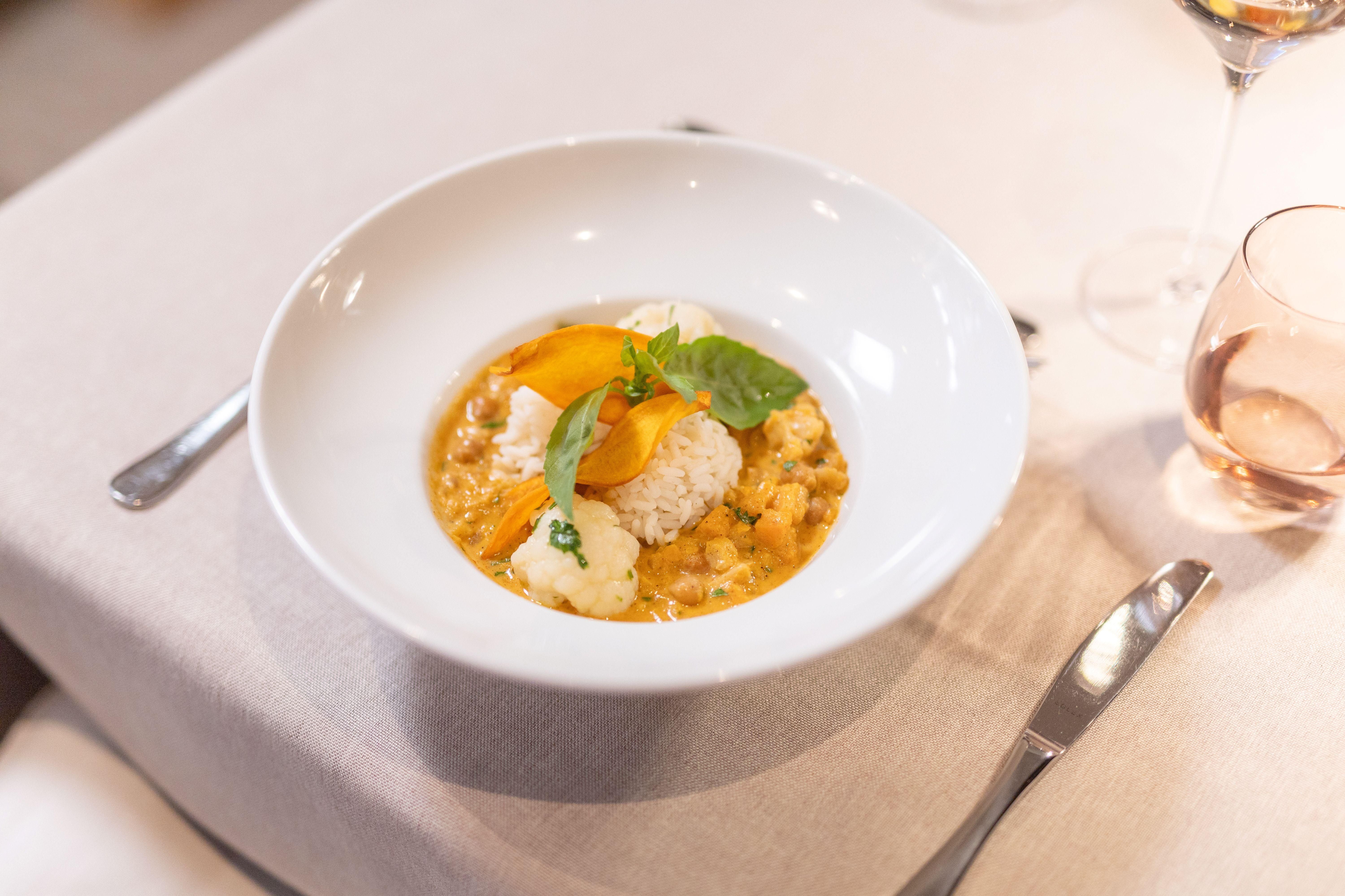 An elegant dish in a white bowl, presented with rice, a creamy sauce, and colorful vegetables. The decoration includes fresh herbs and edible flowers.