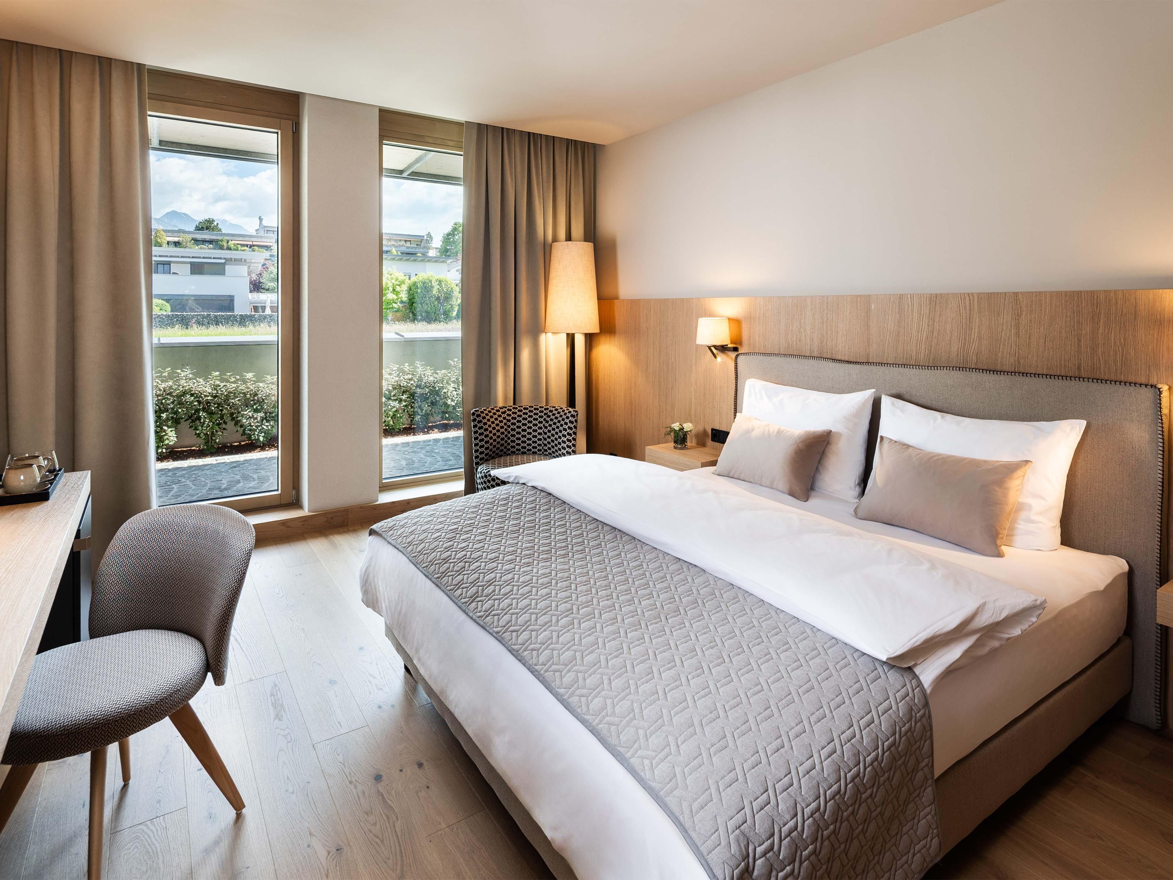 A modern hotel room with a comfortable double bed and a desk. Large windows offer views of the surroundings and let in plenty of daylight.