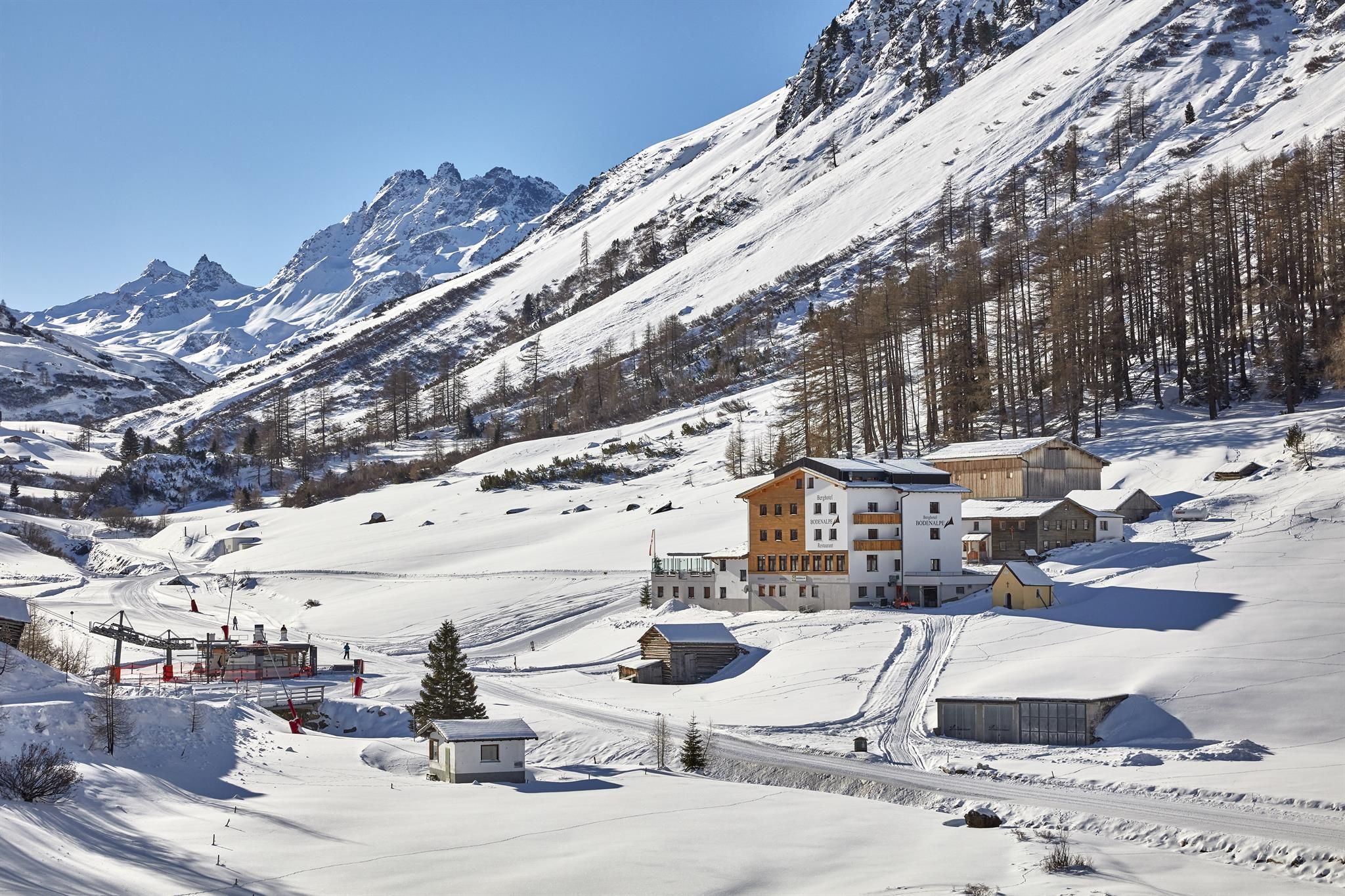A picturesque winter landscape with snow-covered mountains. In the foreground, several buildings are situated in an idyllic, snowy setting.