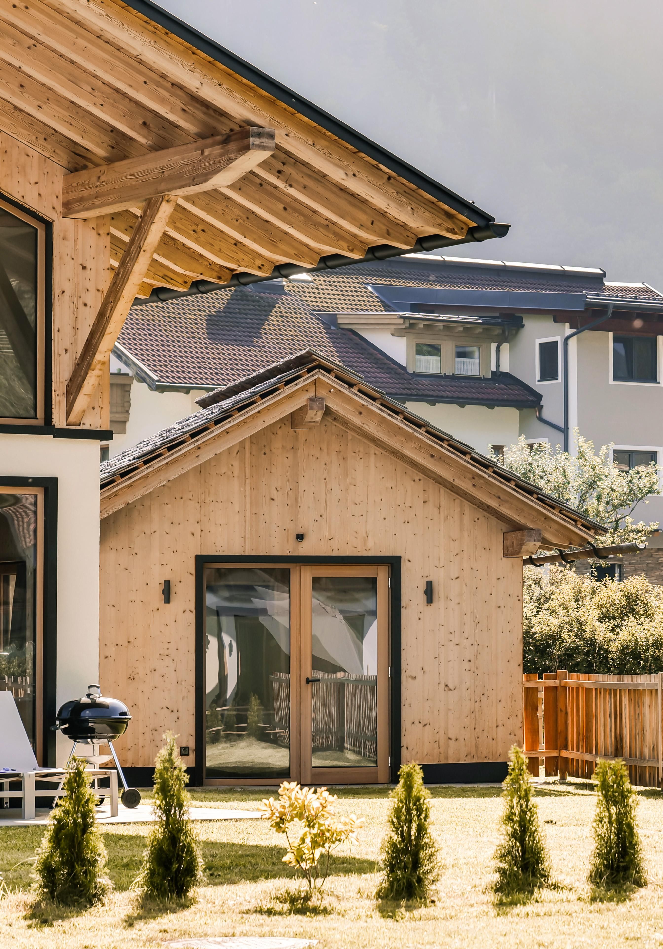 A modern wooden house with an adjacent small building in a well-kept garden. Tall trees and a clear environment surround the architecture.